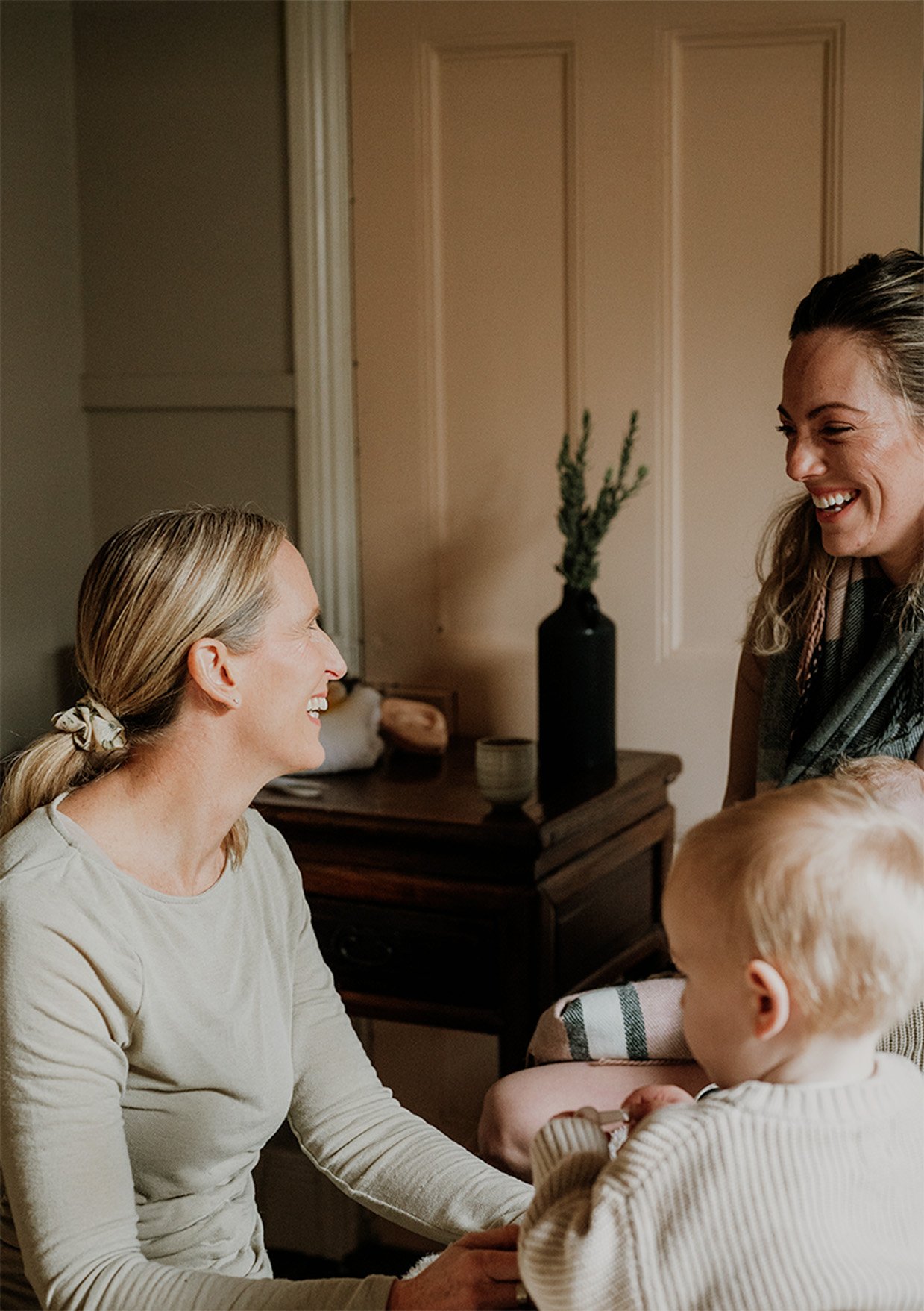 Two women, one with blonde hair in a ponytail and the other with light brown hair, are smiling and interacting with a small child in a cozy, well-lit room. The women are facing each other, and the child is holding something, possibly a toy or food item.