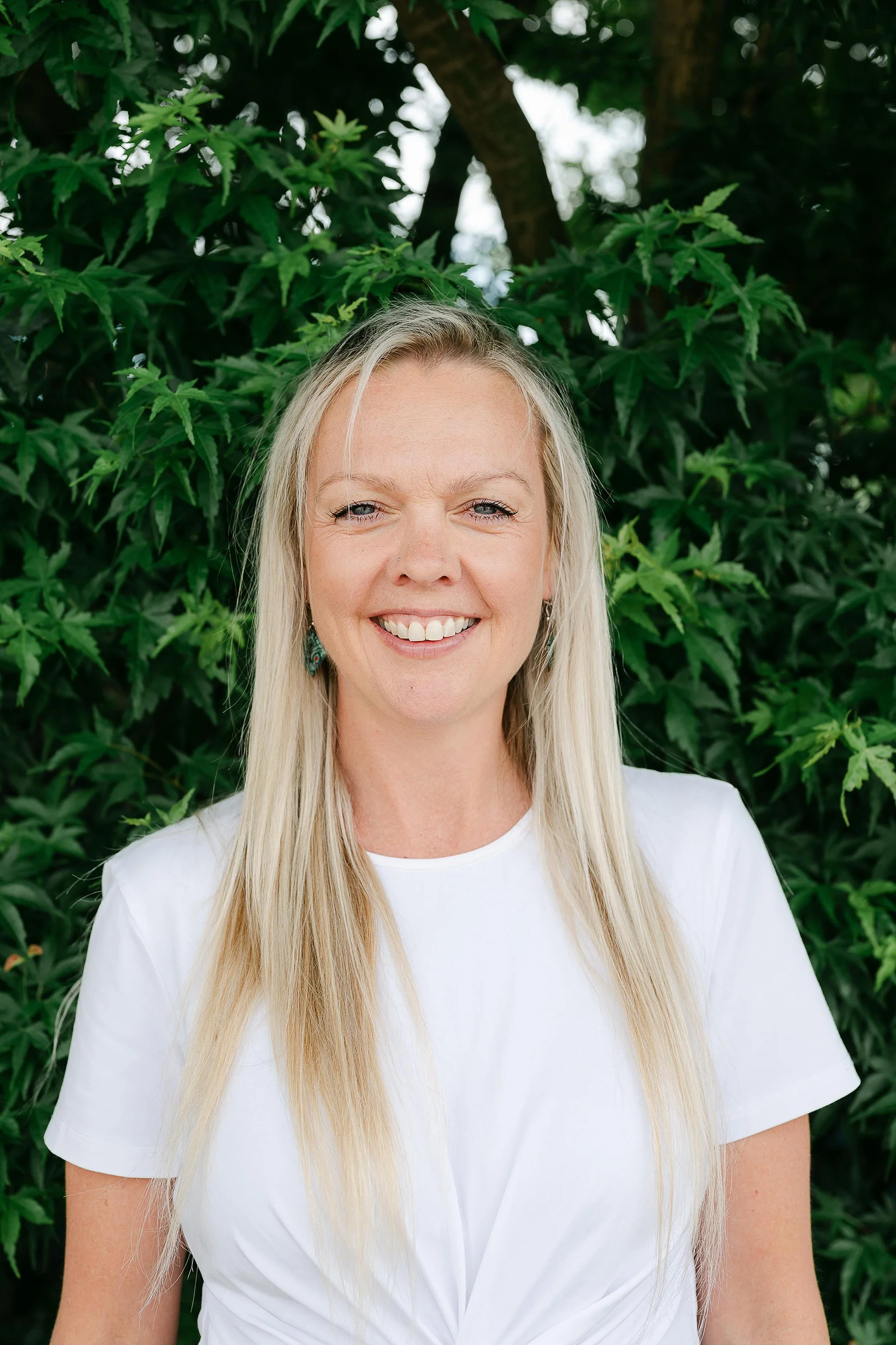 A smiling woman with long blonde hair wearing a white shirt, standing outdoors in front of green foliage.