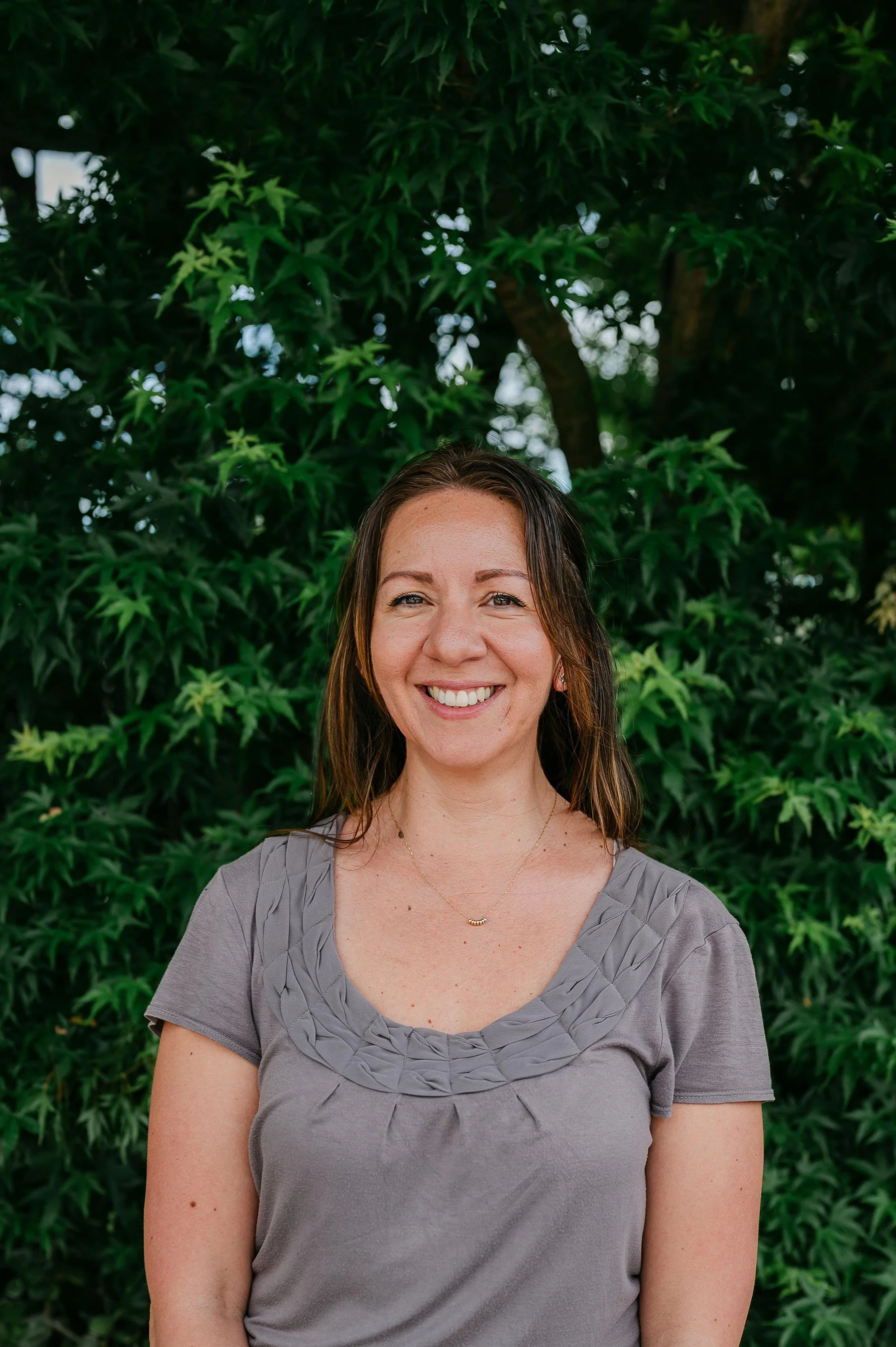 A woman with long brown hair smiling outdoors in front of lush green foliage
