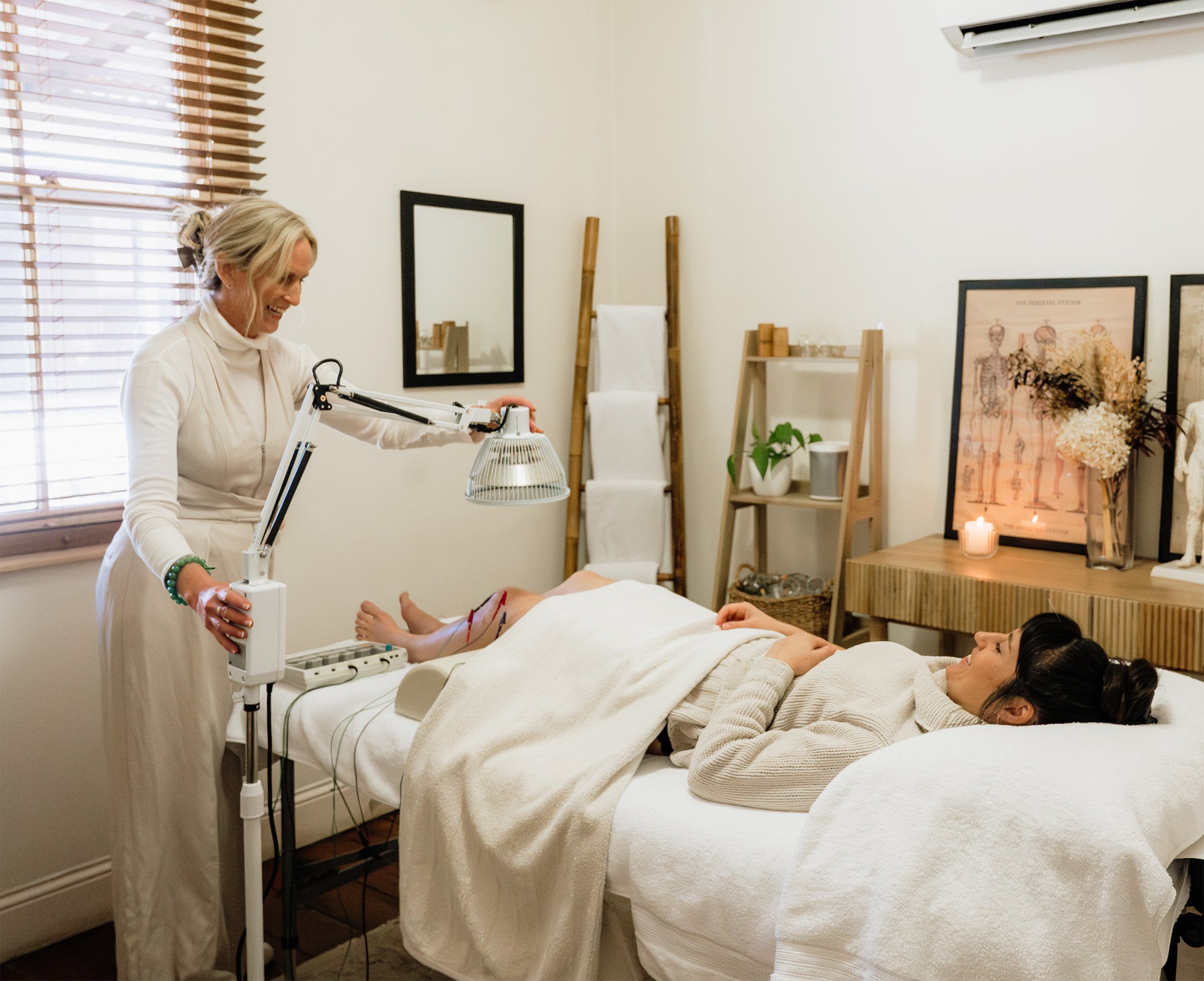 A woman lying on a massage table covered with a white towel having an acupuncture session, with an acupuncturist standing nearby adjusting a lamp.