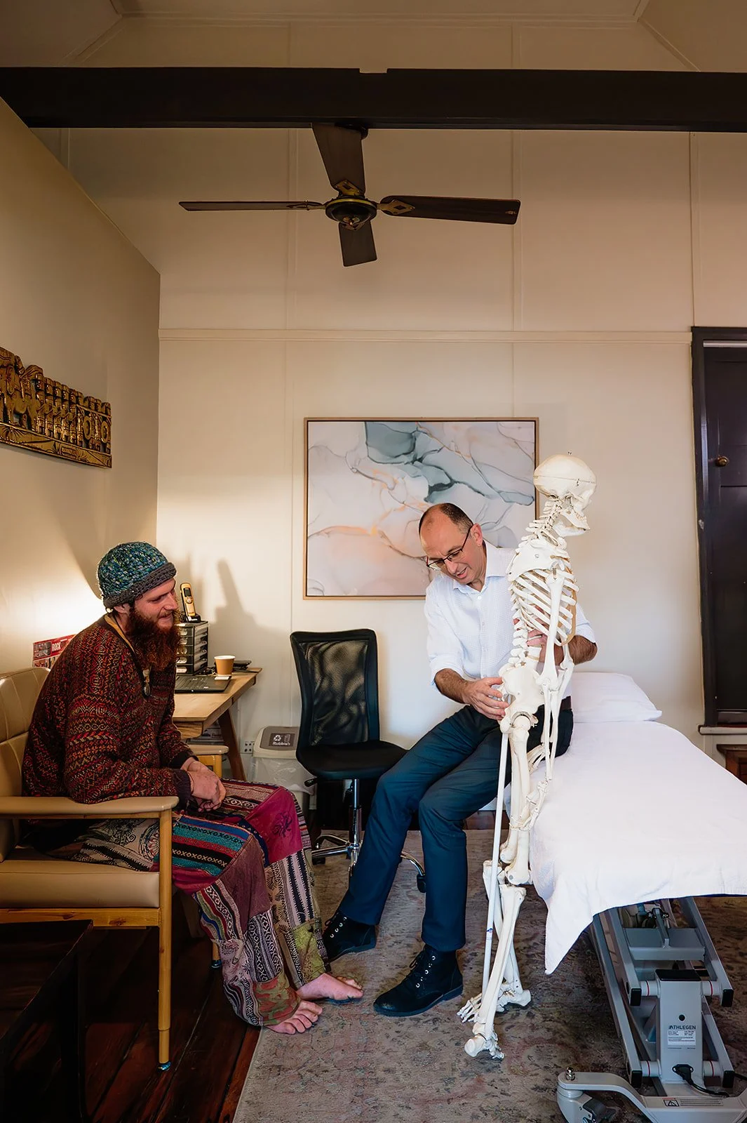 A man sitting on a bed and a woman sitting on a chair having a conversation with a life-sized skeleton model in a medical office or clinic.