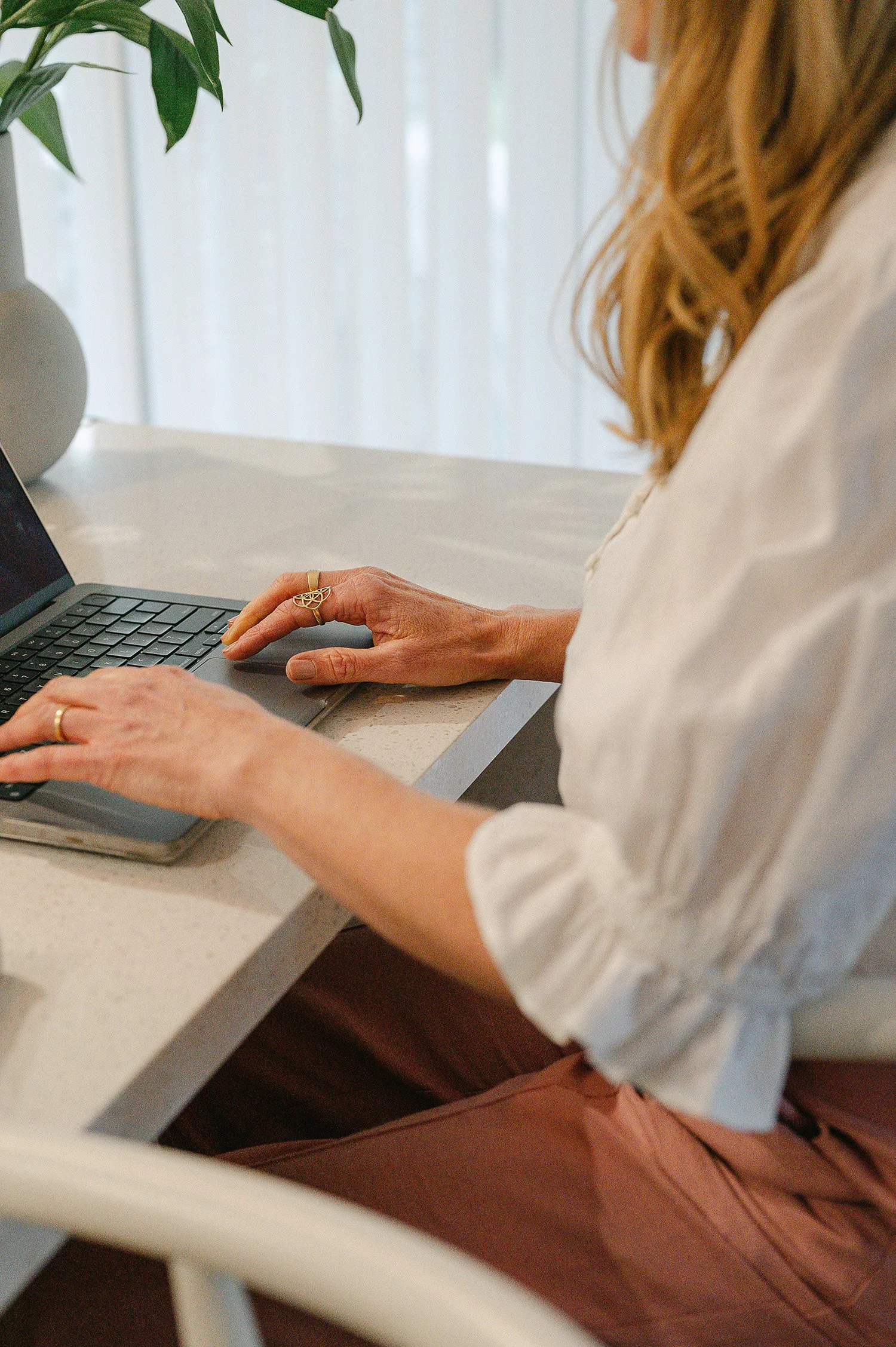 A woman with wavy, shoulder-length hair, wearing a white shirt with rolled-up sleeves, is typing on a laptop at a white table near a window with sheer curtains and a decorative plant in a white vase.