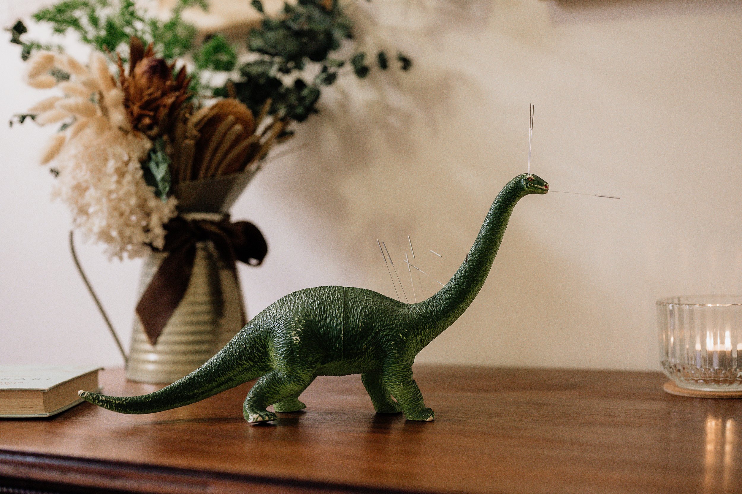 A green plastic dinosaur toy with thin wires protruding from its head and back, sitting on a wooden surface next to a book and a glass candle holder, with a vase of dried and fresh flowers in the background.