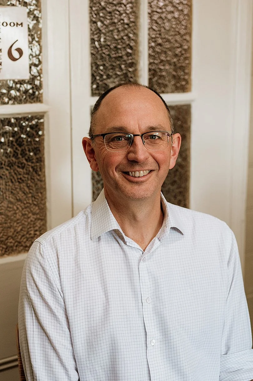 A smiling man wearing glasses and a white checkered shirt sitting indoors in front of a white wall with decorative glass panels.
