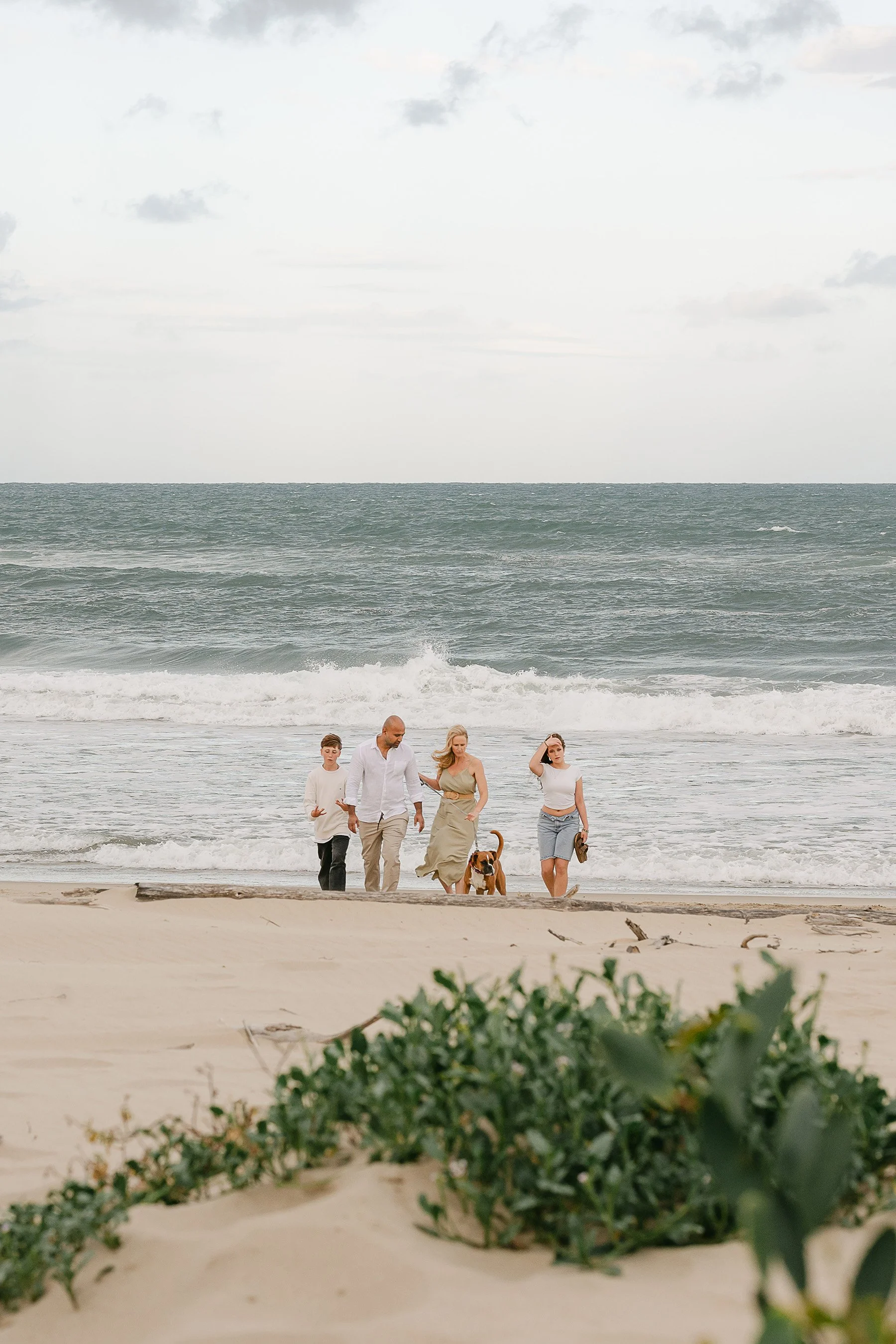 A family walking along the beach with their dog, with the ocean and sky in the background.