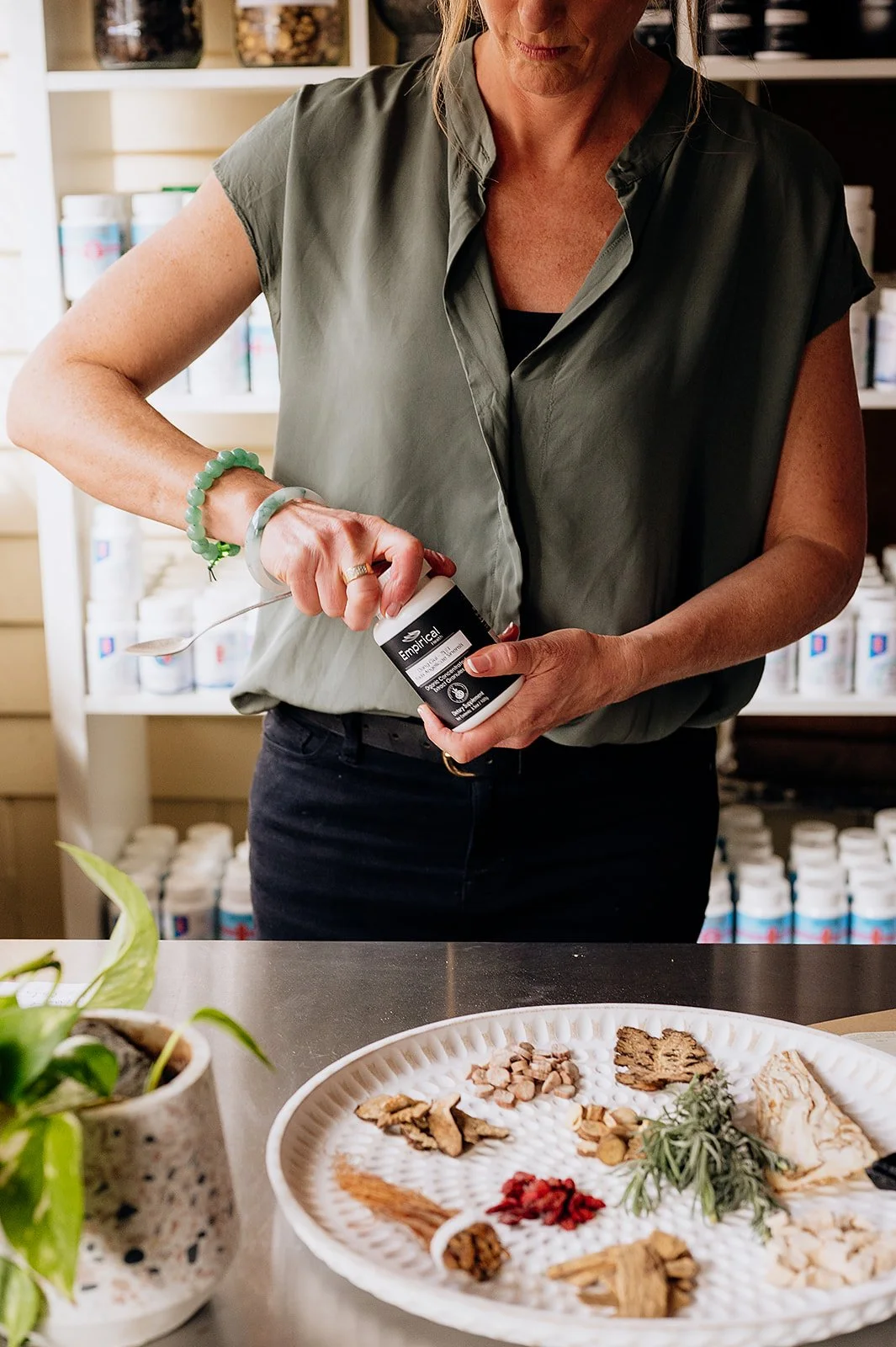 A woman opening a supplement container at a table with herbs and dried flowers, with shelves of bottles in the background.
