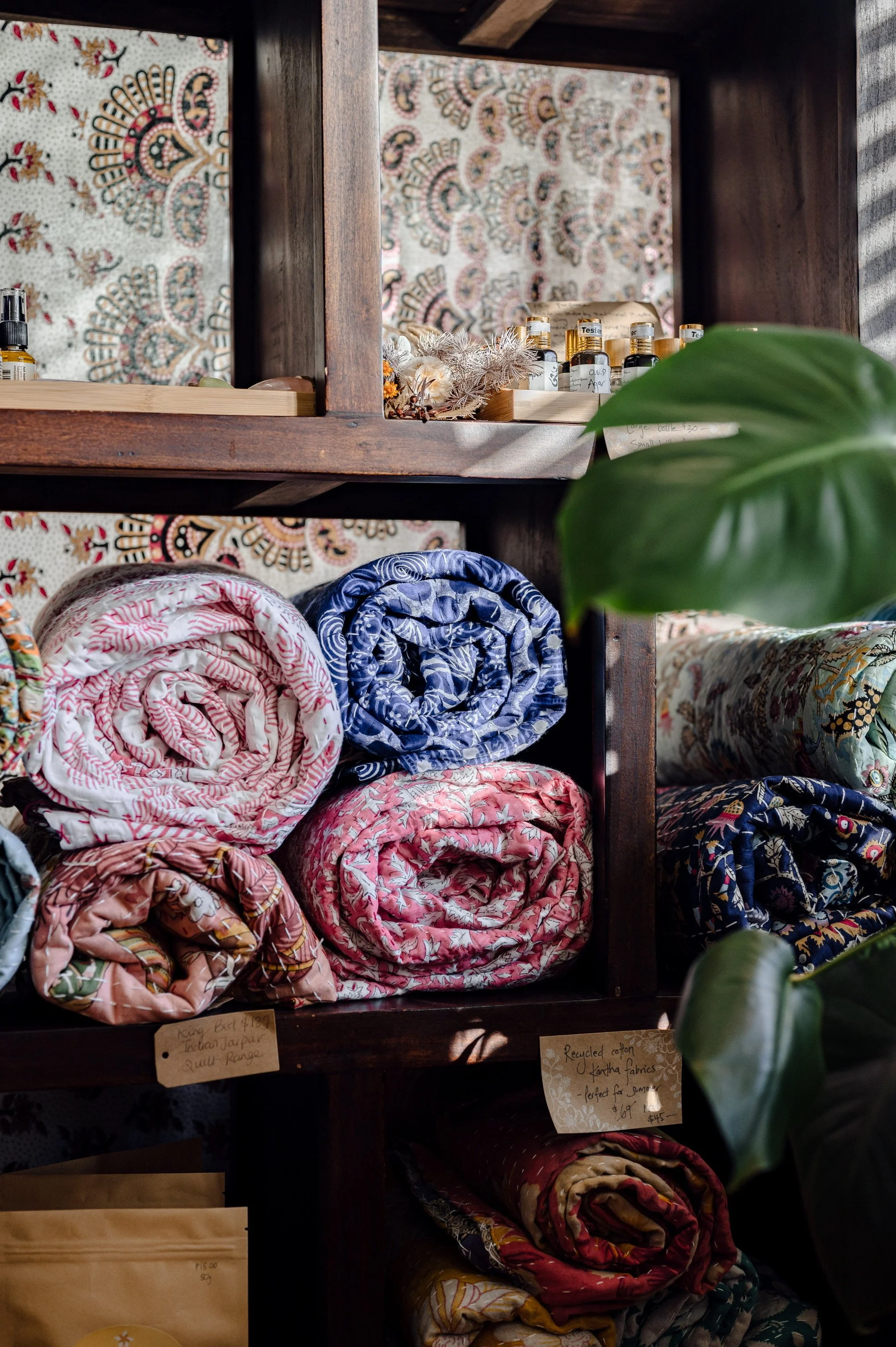 Folded colorful patterned fabric and quilted blankets on a dark wooden shelf inside a store, with tags and labels visible.
