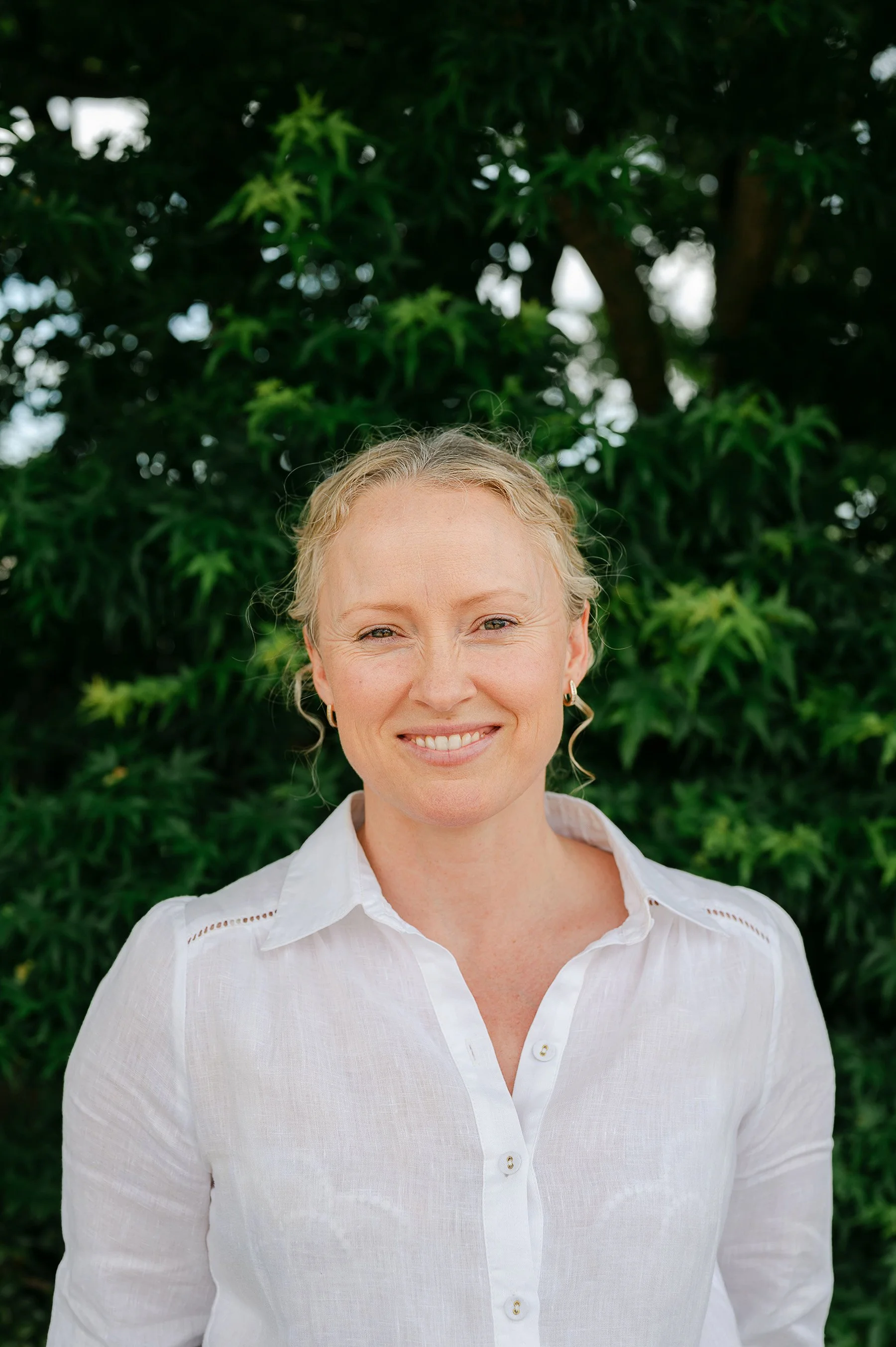 Portrait of a woman with blonde hair, smiling, wearing a white shirt, standing outdoors with green foliage in the background.