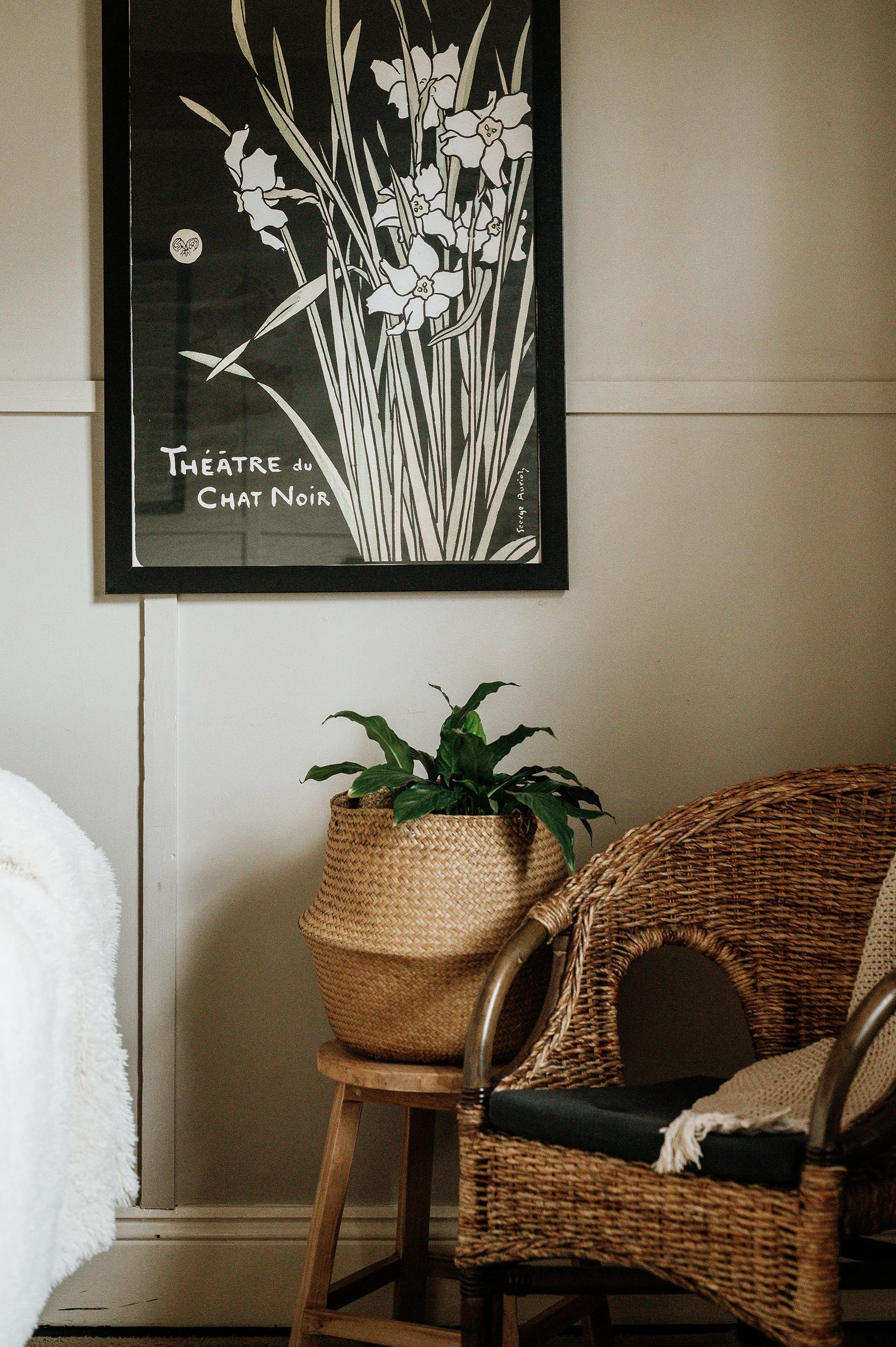 Interior corner with rattan chair, potted plant in woven basket on a small wooden stool, and framed black-and-white poster on the wall reading 'Théâtre du Chat Noir'.