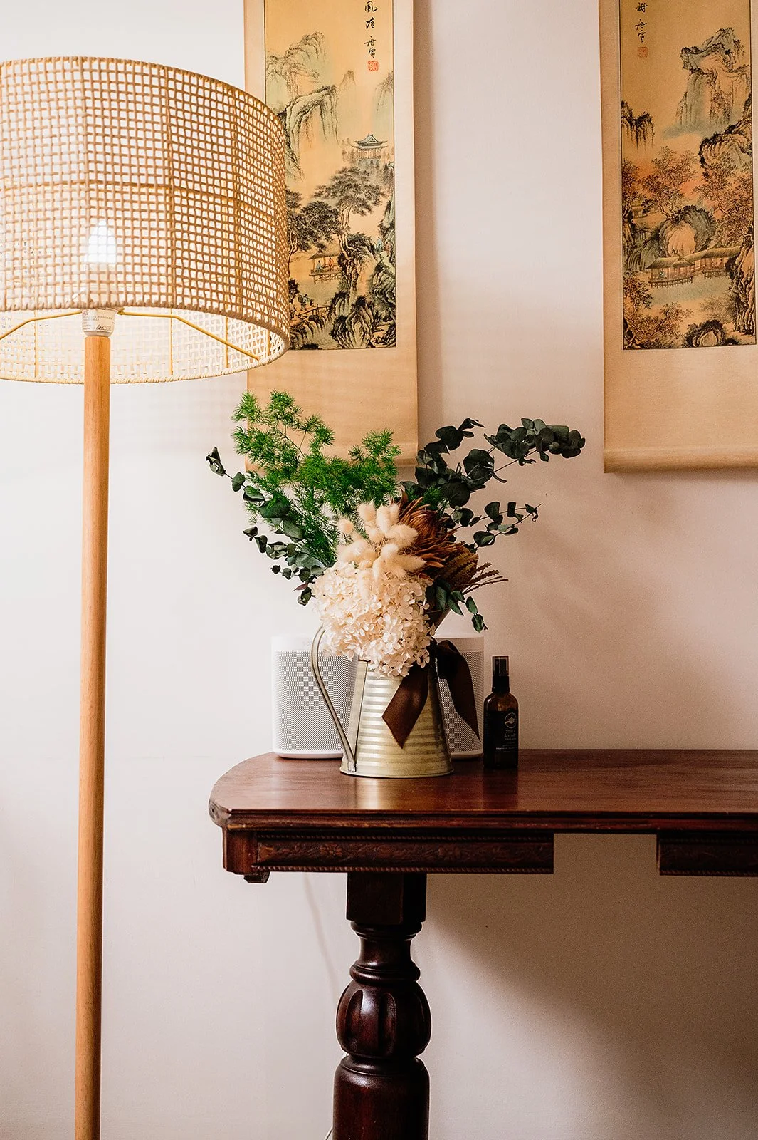 A wooden table with a metal vase of flowers, a small speaker, and a small bottle, beside a standing lamp with a woven shade, against a wall decorated with traditional Asian-style landscape paintings.