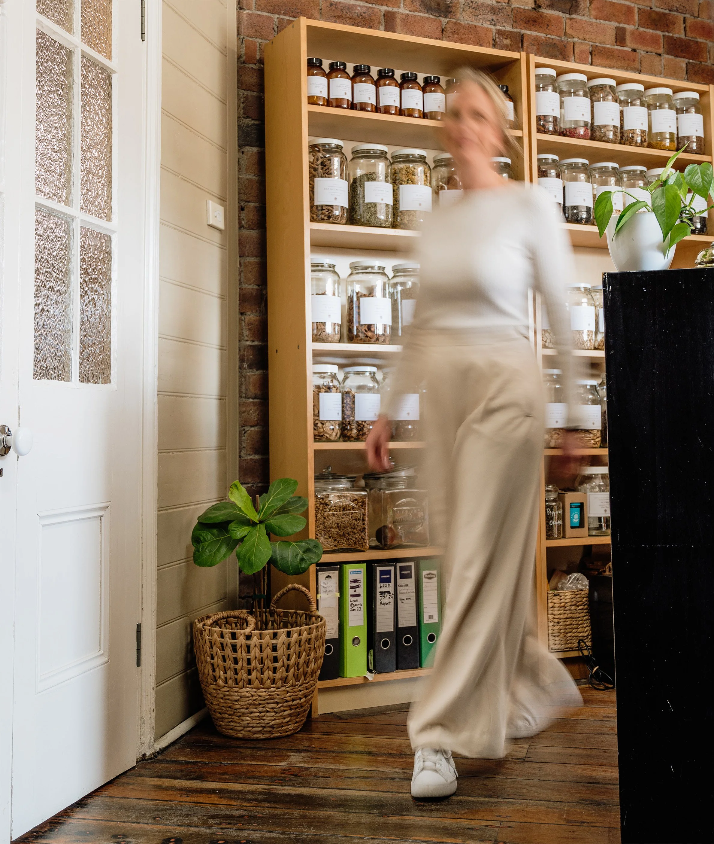 A blurred woman in beige pants and shoes walking past glass jars of dried herbs and spices on a wooden shelf in a cozy room with a brick wall and wooden floor, next to a potted plant in a woven basket.