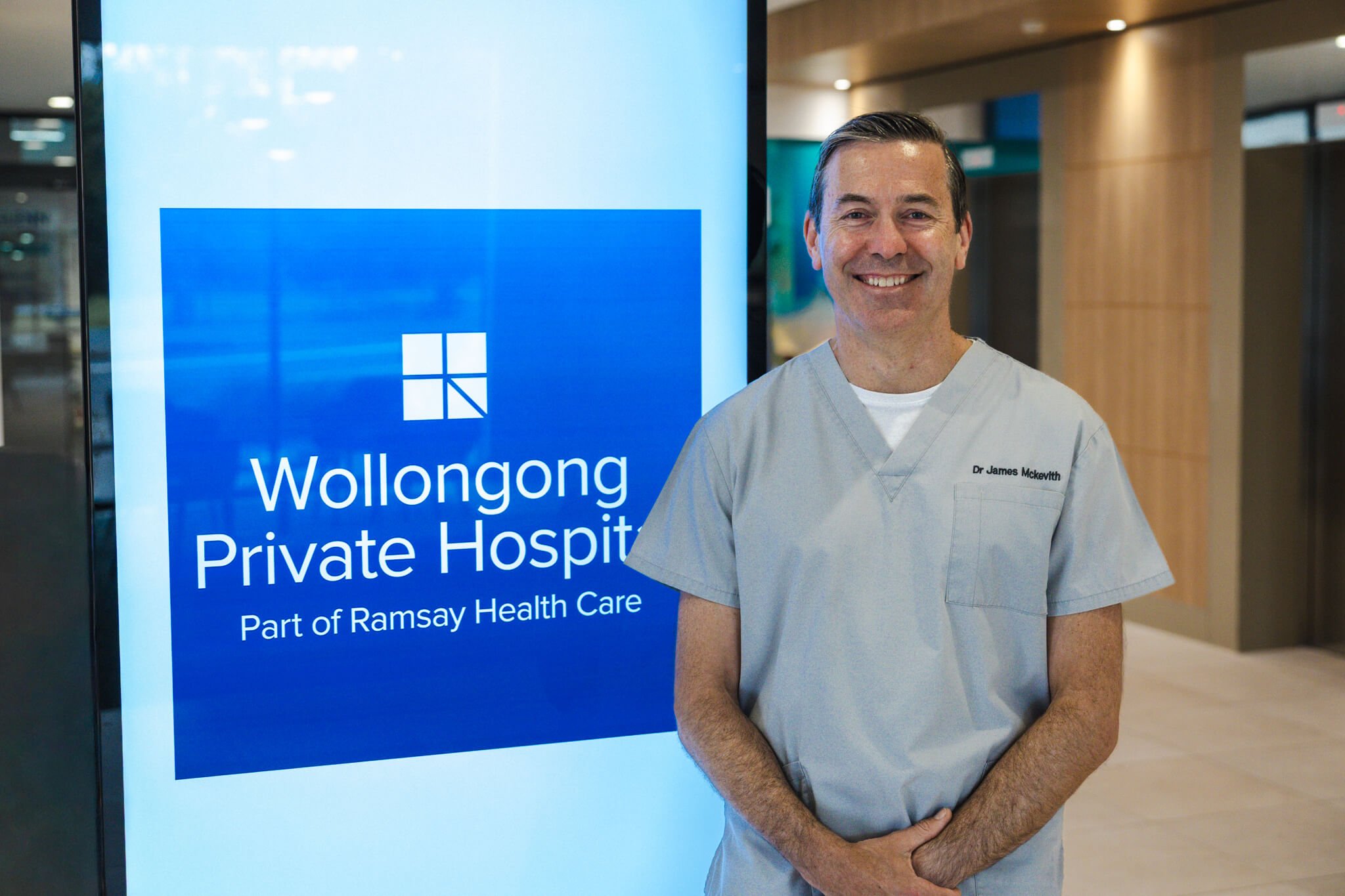 A smiling male doctor in scrubs standing next to a digital sign displaying 'Wollongong Private Hospital, Part of Ramsay Health Care' at a hospital lobby.