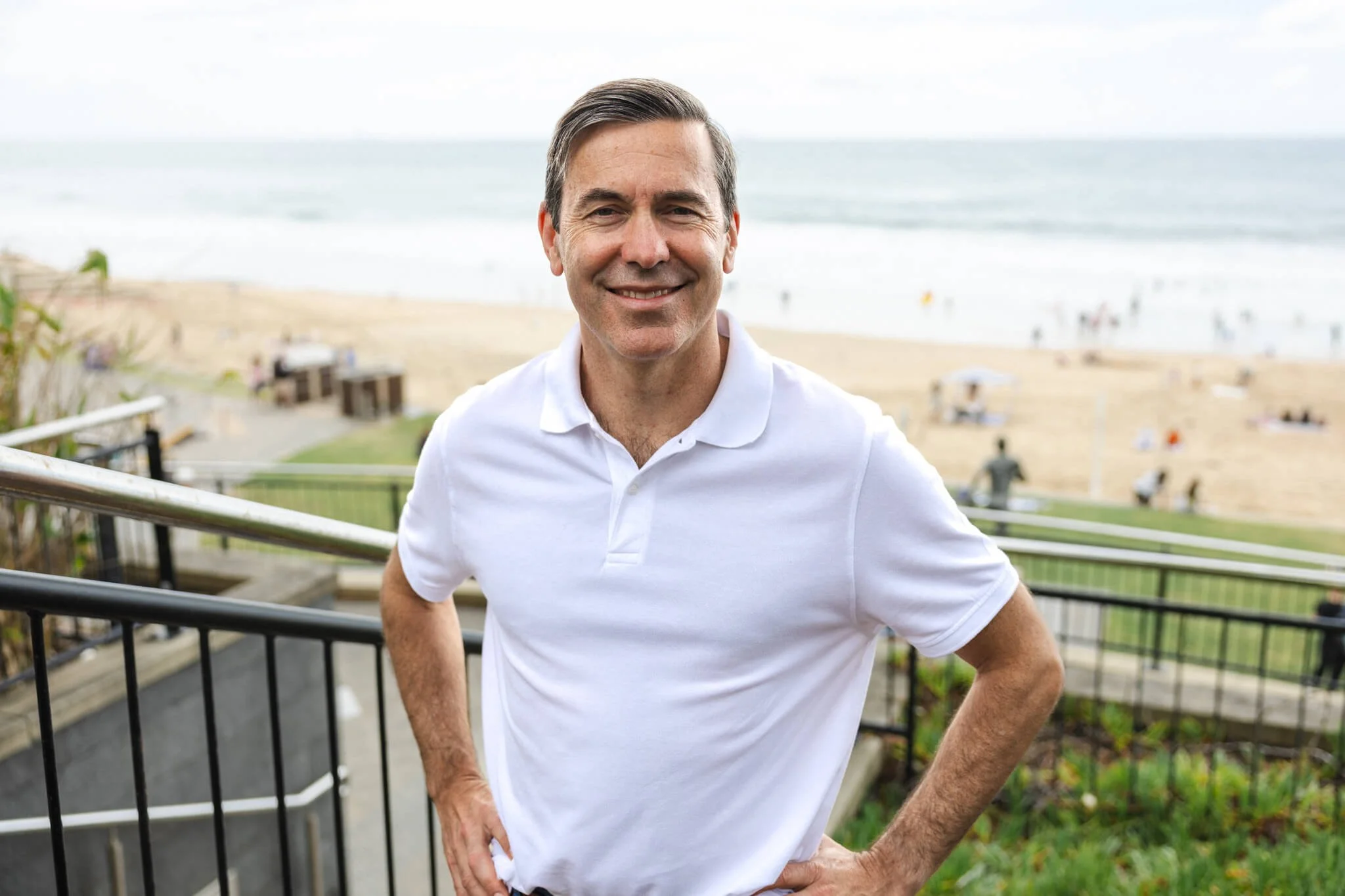 A man in a white polo shirt smiling at the camera, standing outdoors on a stairway near a beach with sand and ocean in the background.
