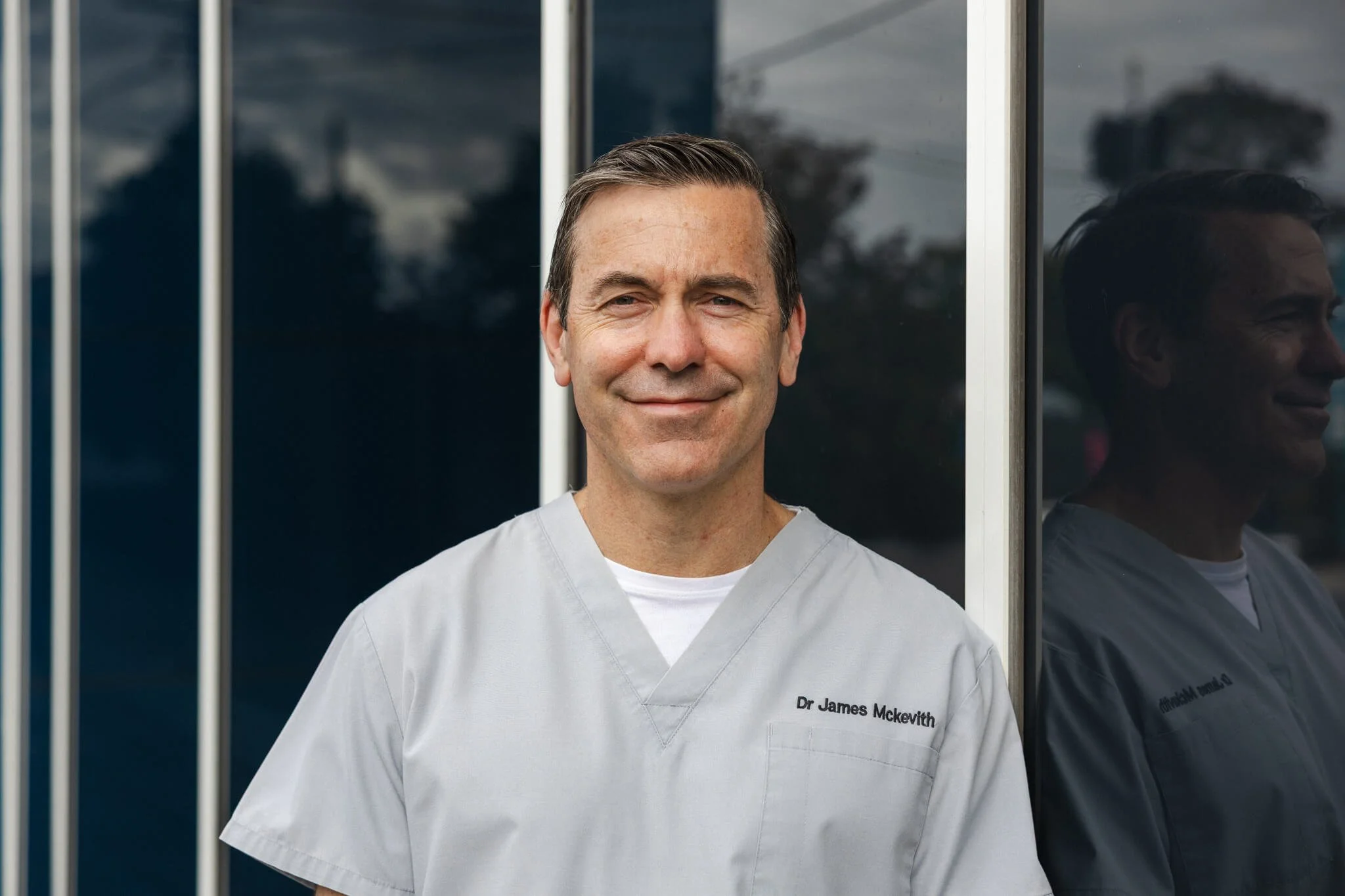 smiling male doctor in white coat with name tag 'Dr James McKevith' standing outside near a building with glass walls.