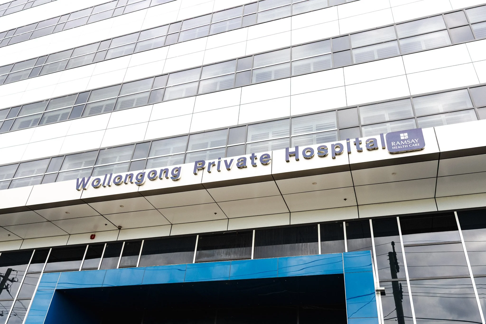 Front of a modern multi-story building with a sign that reads 'Wollongong Private Hospital' and a smaller sign for 'Ramsay Health Care'.