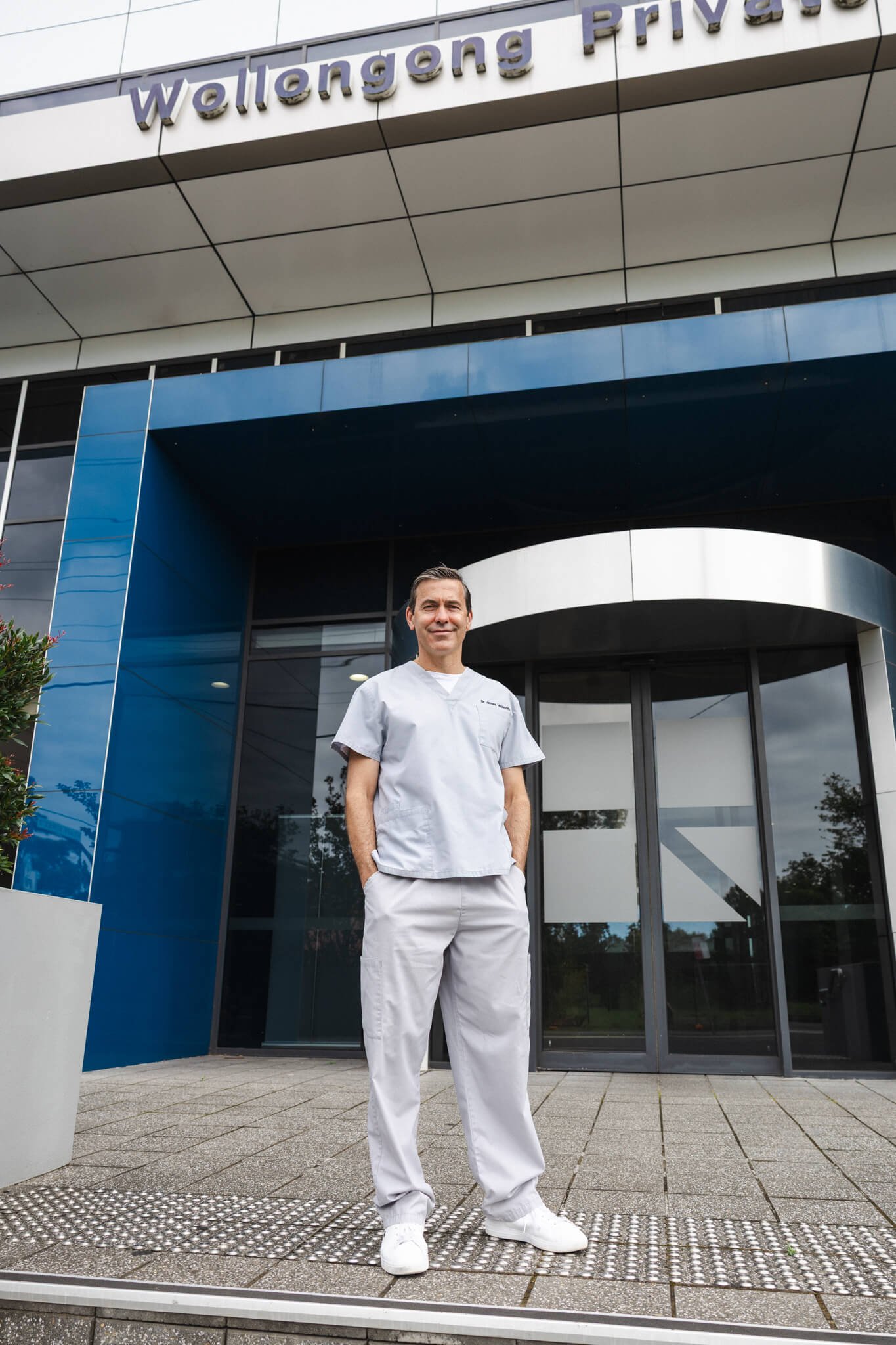 A smiling male healthcare professional standing outside the Wallong Private building, wearing light gray scrubs and white shoes, near the entrance with glass doors and modern architecture.