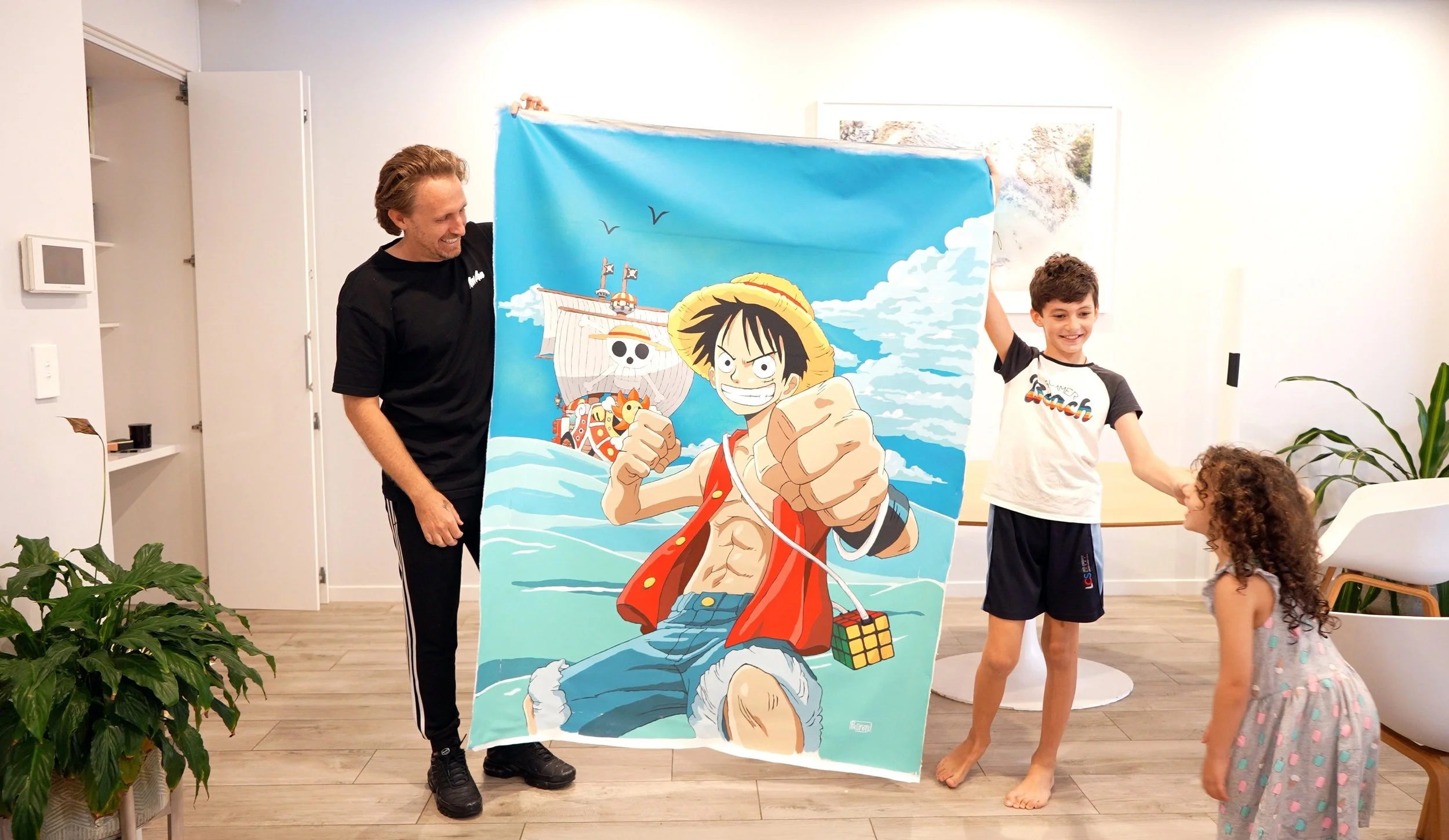 Three people in a living room holding up a large, colorful poster of Monkey D. Luffy from the anime One Piece. The poster shows Luffy with a straw hat, smiling and pointing forward with a ship in the background.
