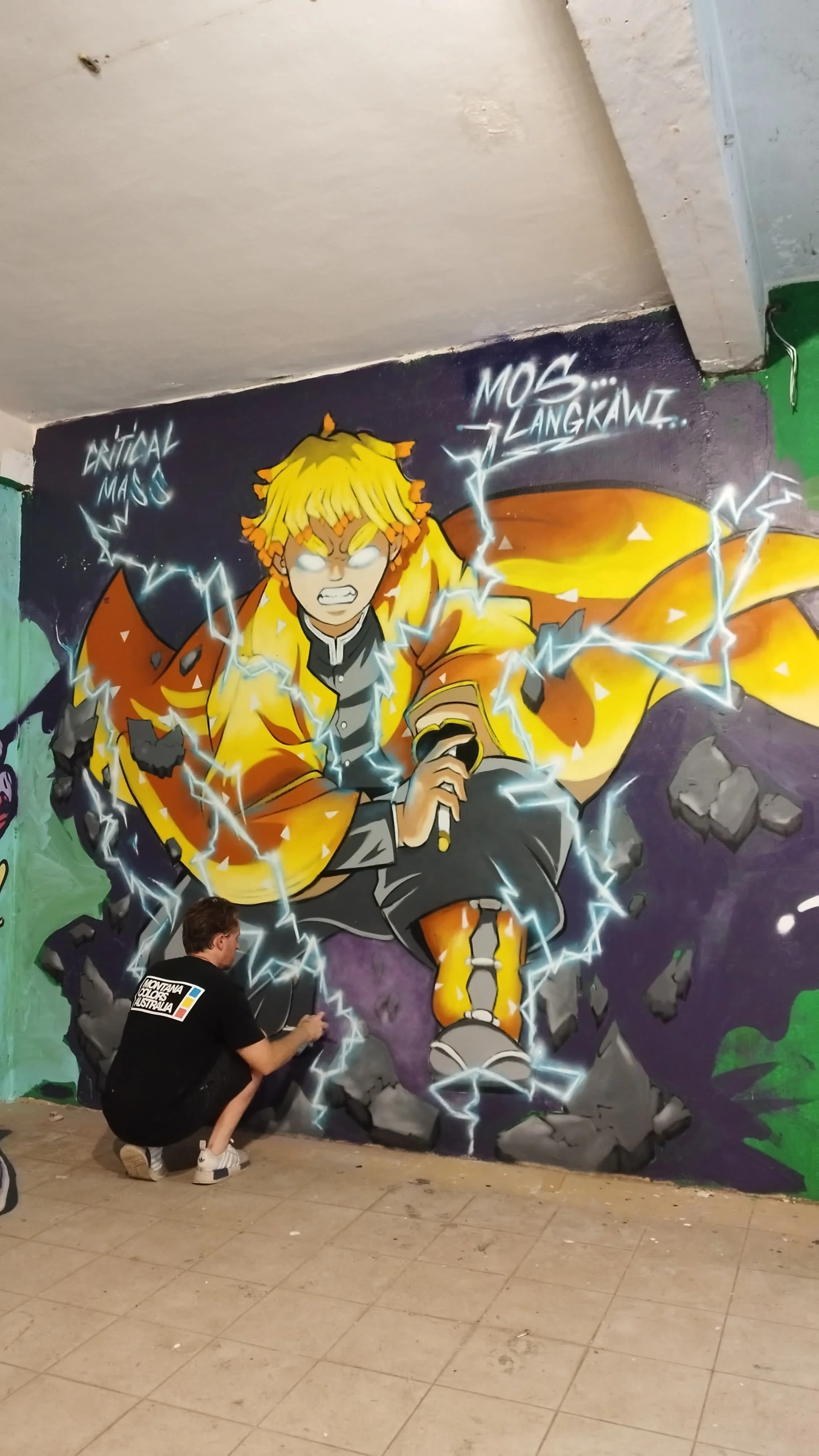 A man is painting a vibrant graffiti mural of a muscular anime character with glowing white eyes, yellow hair, and an orange jacket, breaking through a wall with lightning effects. The wall has words like 'Critical Mass' and 'Mos... Lankawi' written above the character.