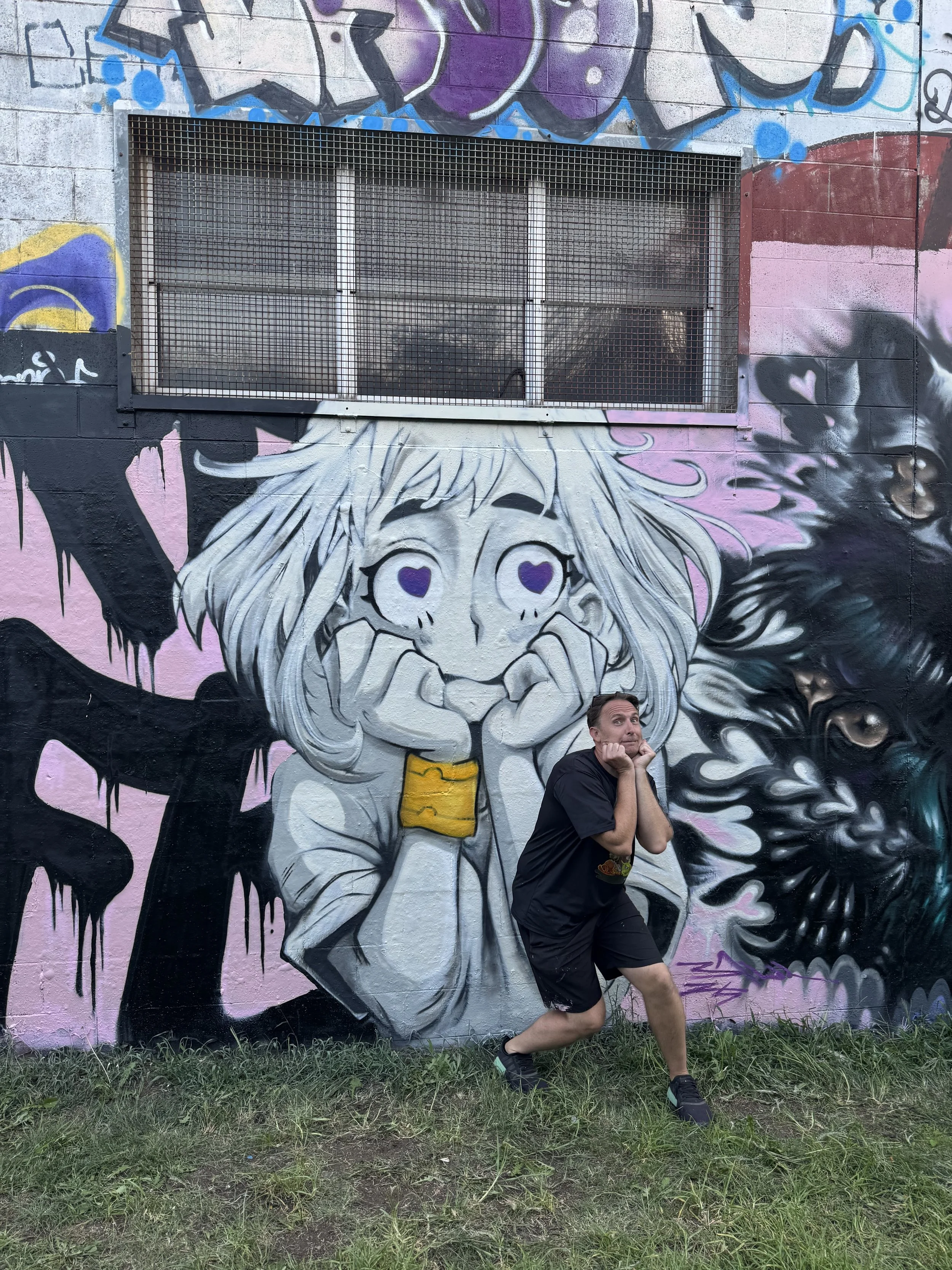 A man making a spooky pose in front of a colorful mural featuring a cartoon girl with heart-shaped eyes and long hair, alongside a black and gray dragon or monster face.