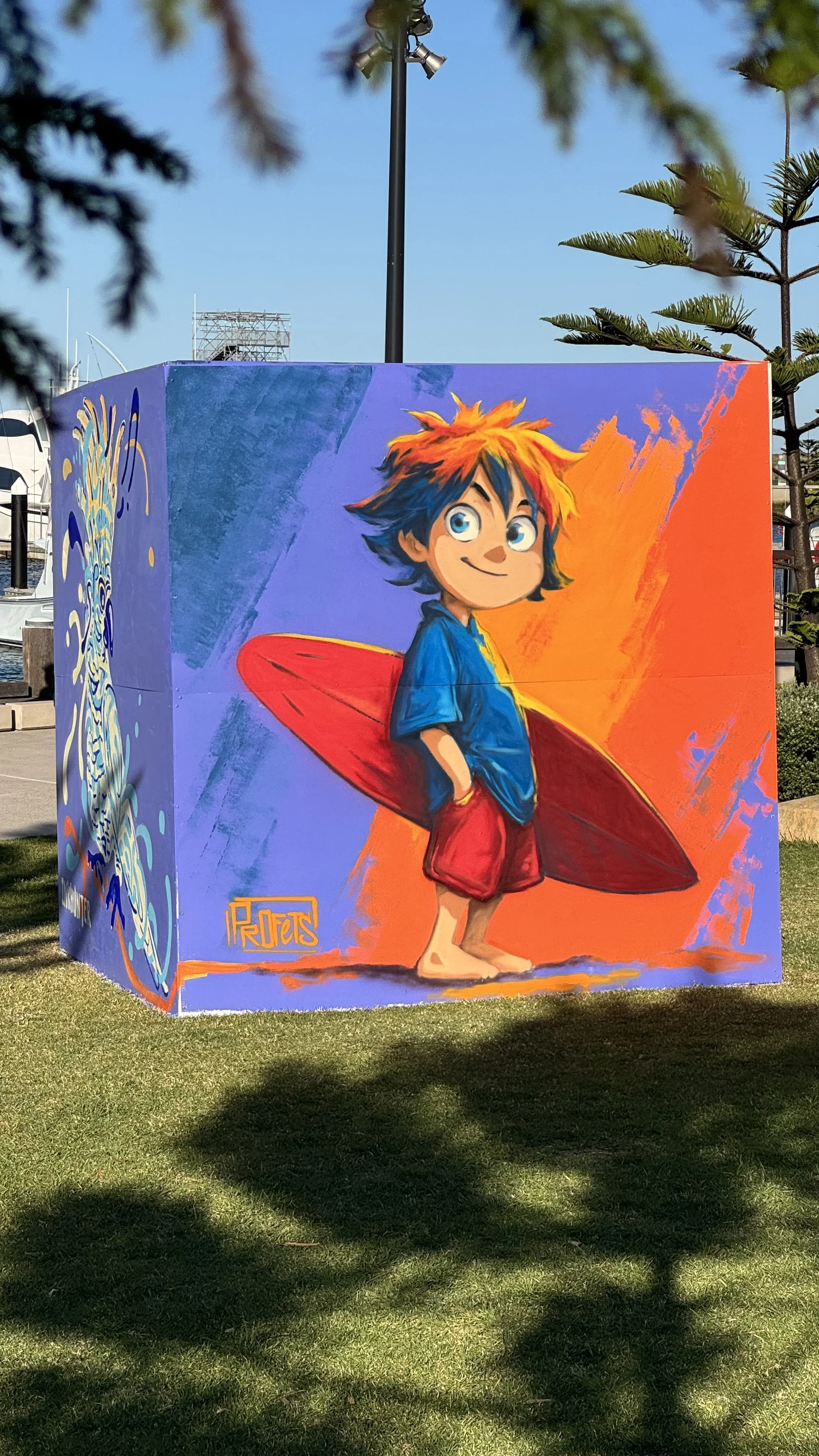 Colorful mural of a young boy with blue eyes, spiky multicolored hair, wearing a blue shirt and red shorts, holding a red surfboard, on an outdoor wall.