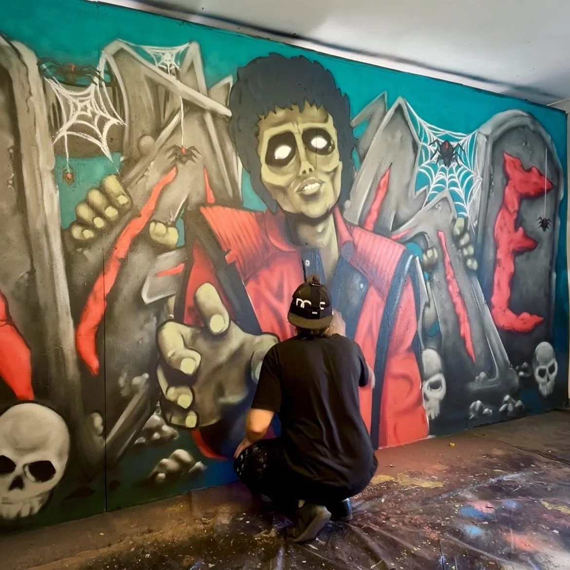 A person with a cap kneeling to paint a graffiti mural of a skeleton figure with skull face, holding a hammer, surrounded by cobwebs, skulls, a red dog, and the word "DIE" in red letters.
