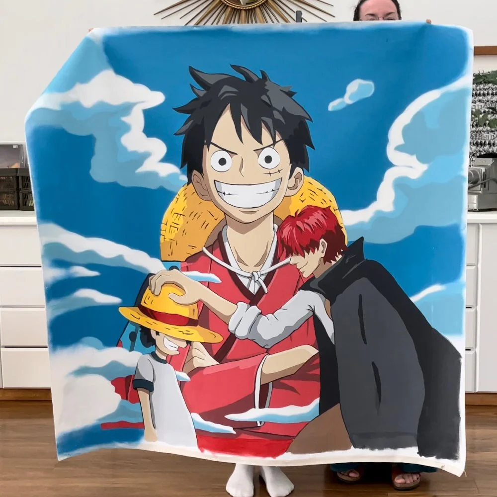 A large blanket with anime characters from One Piece, including Monkey D. Luffy with a straw hat, smiling broadly, and two other characters, one with red hair and a black cloak, and a young boy with a yellow hat and gray shirt. The background features a sky with clouds.