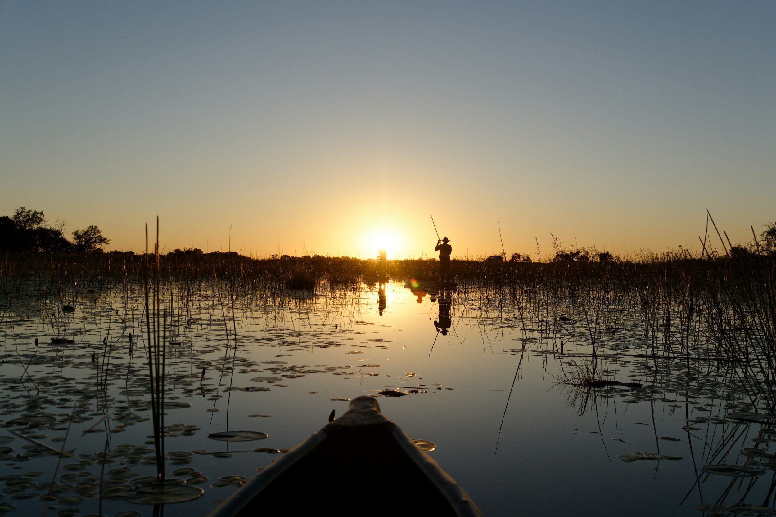 A silhouette of two people fishing on a lake at sunset, seen from the front of a boat with lily pads on the water.