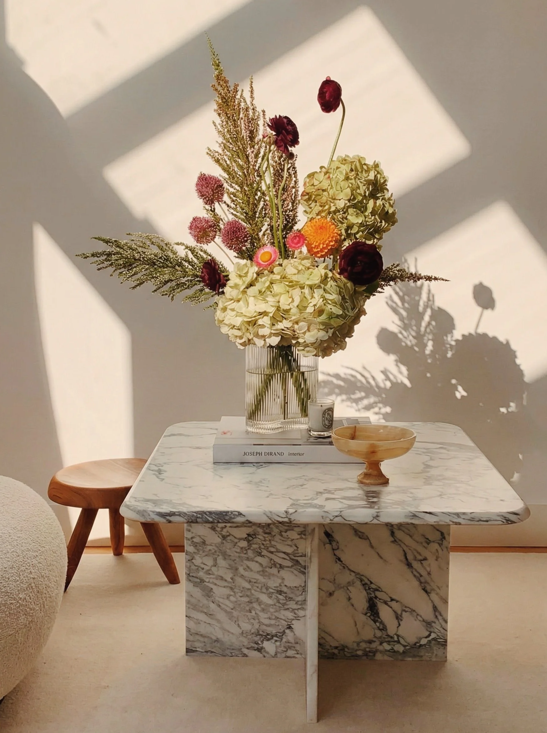 A marble coffee table with a large floral arrangement in a clear glass vase, a small wooden bowl, and a book titled 'Joseph Dirand Interior'. The table is on a beige carpet, with sunlight casting shadows on the white wall behind.