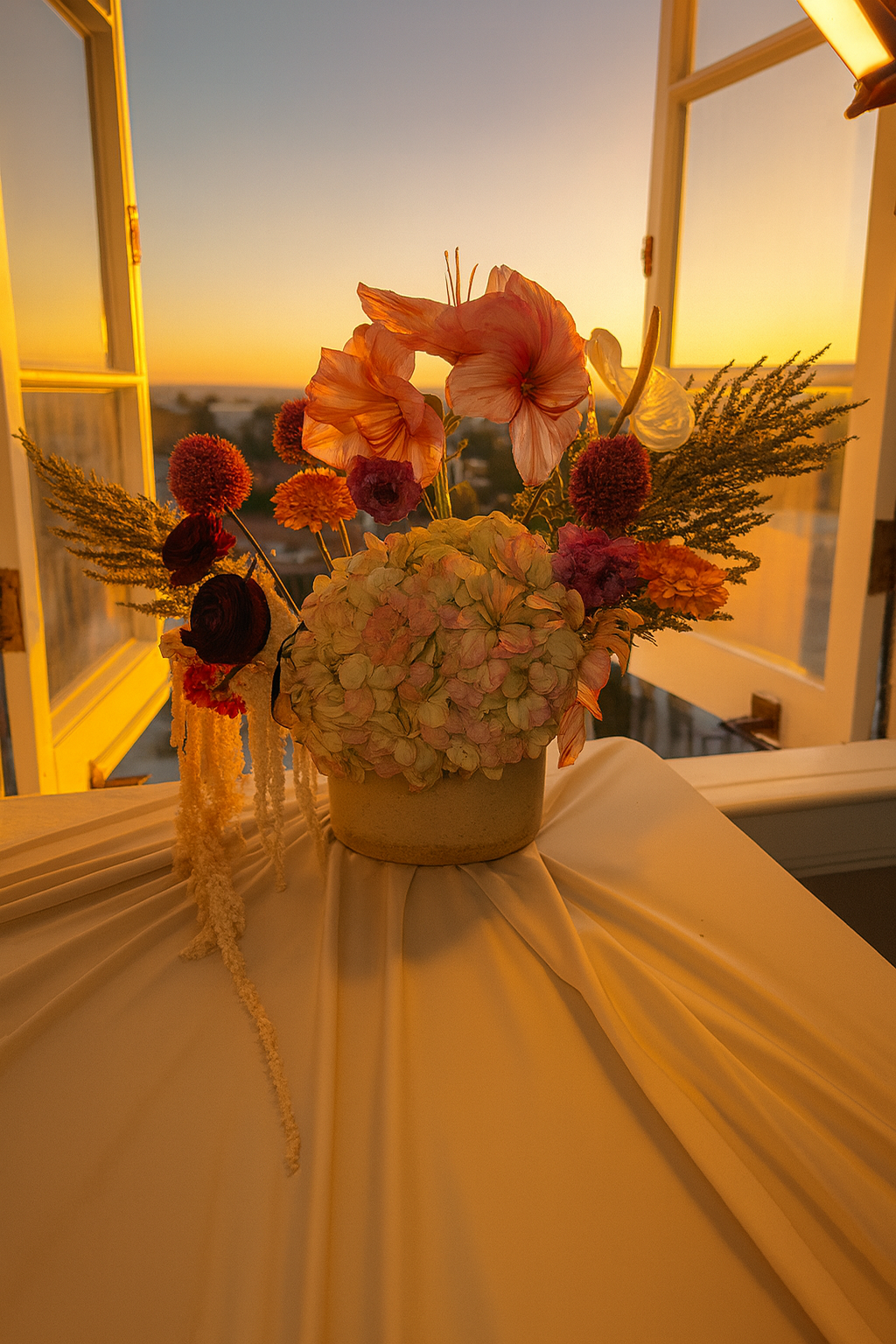 A flower arrangement with pink, red, and orange flowers in a beige vase on a table with draped white cloth, against a sunset view through open windows.