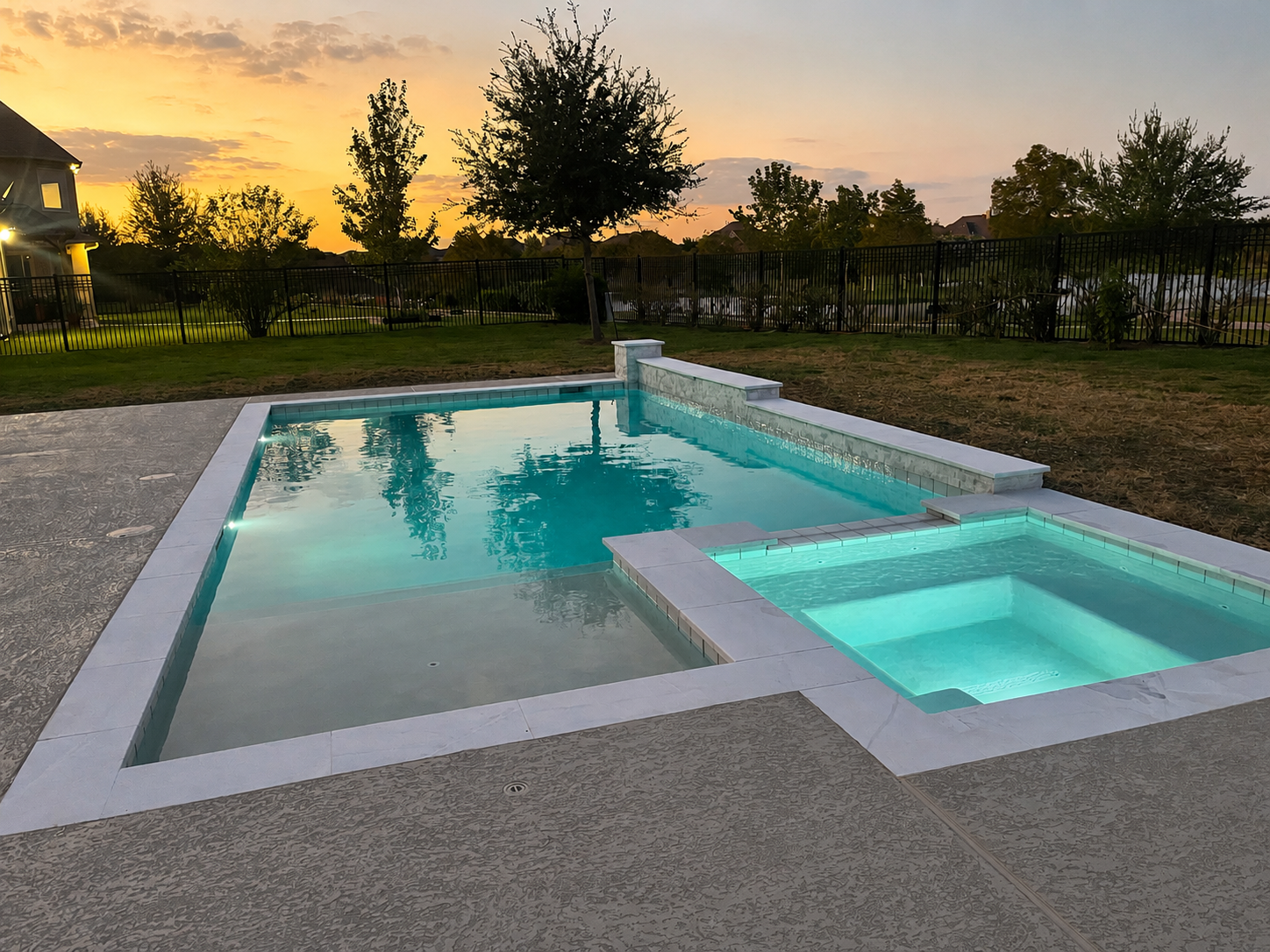 A backyard with a swimming pool and a hot tub at sunset. The pool has built-in lighting, and the yard is enclosed by a black metal fence. There are trees and a house in the background, with a partly cloudy sky.