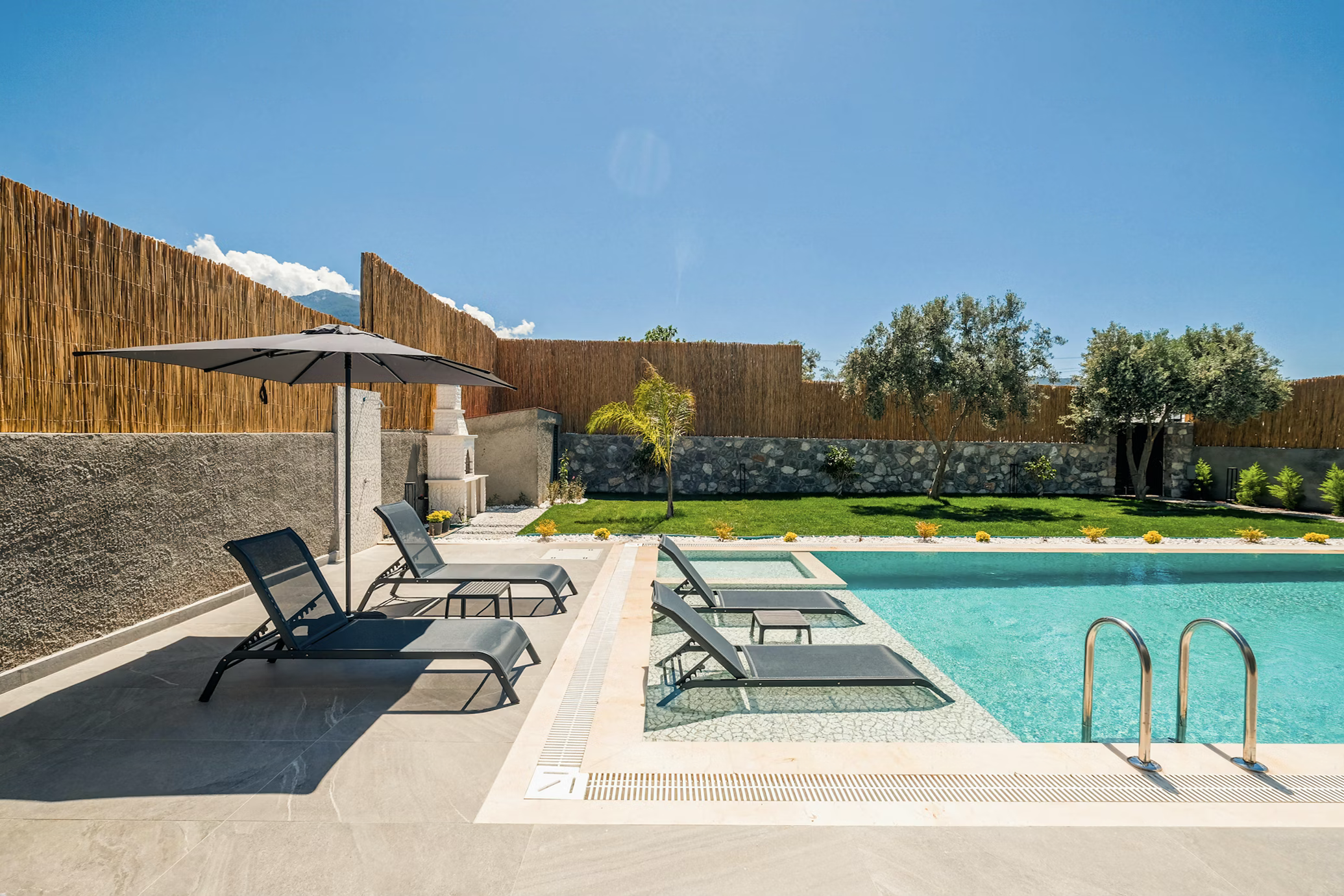 A backyard swimming pool with lounge chairs, an umbrella, and a landscaped yard enclosed by a wooden fence.
