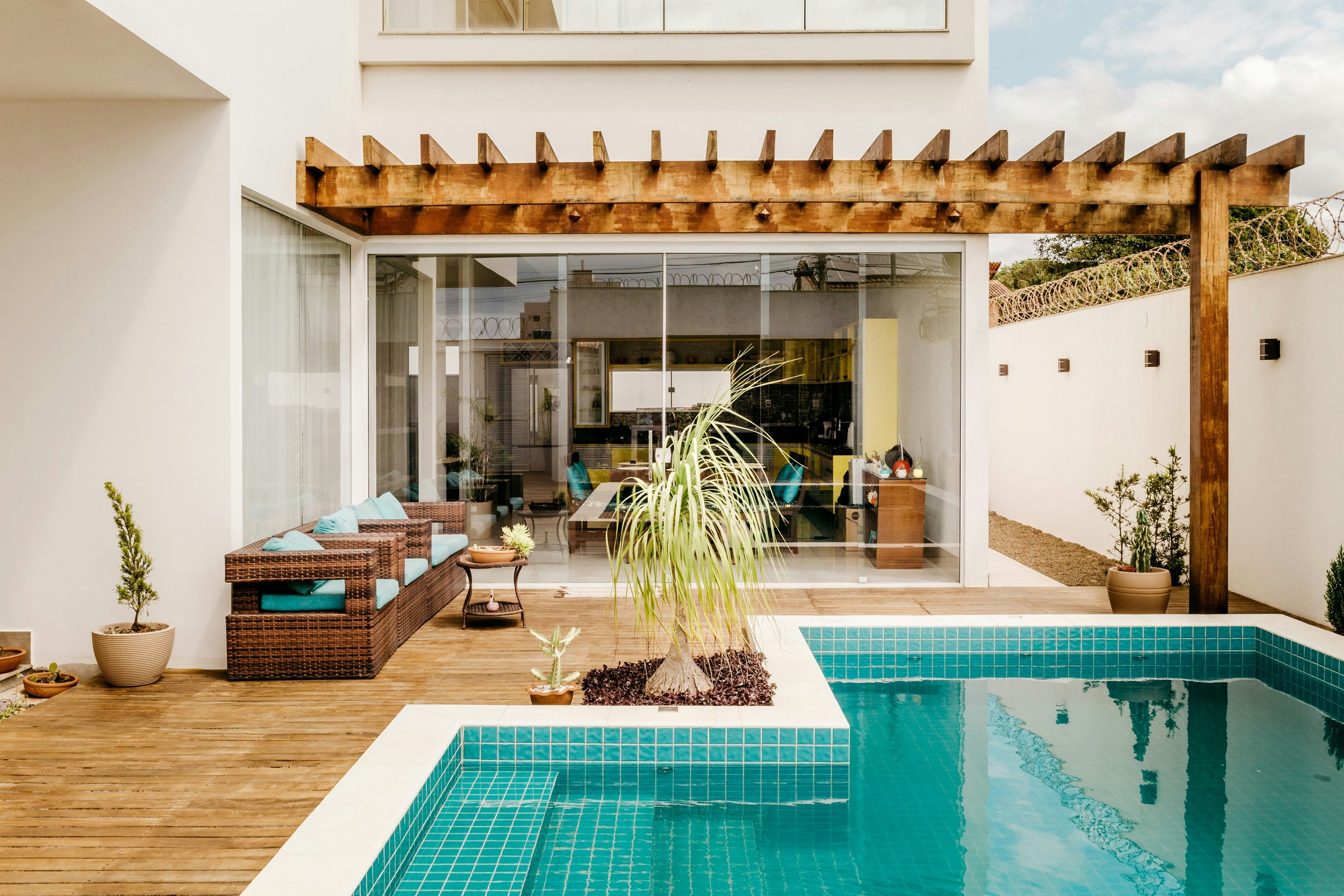 Modern house backyard with swimming pool, wooden deck, outdoor seating, potted plants, and glass sliding doors leading inside.