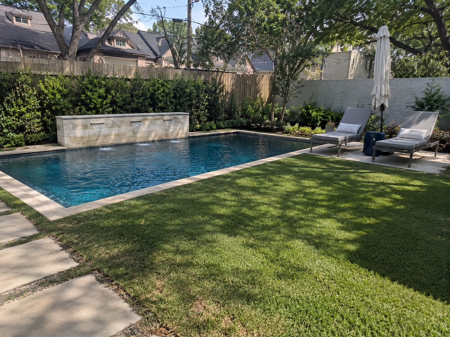 A backyard with a rectangular swimming pool, surrounded by green grass and greenery. There are two lounge chairs with cushions, a closed umbrella, and a water feature with three waterfalls on a stone wall.