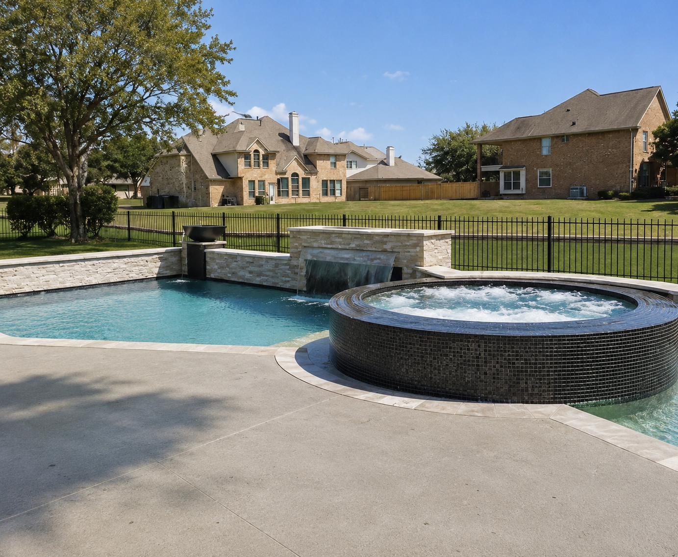 Residential backyard with a swimming pool and hot tub, enclosed by a black metal fence, with houses and trees in the background on a clear sunny day. Humble, Tx