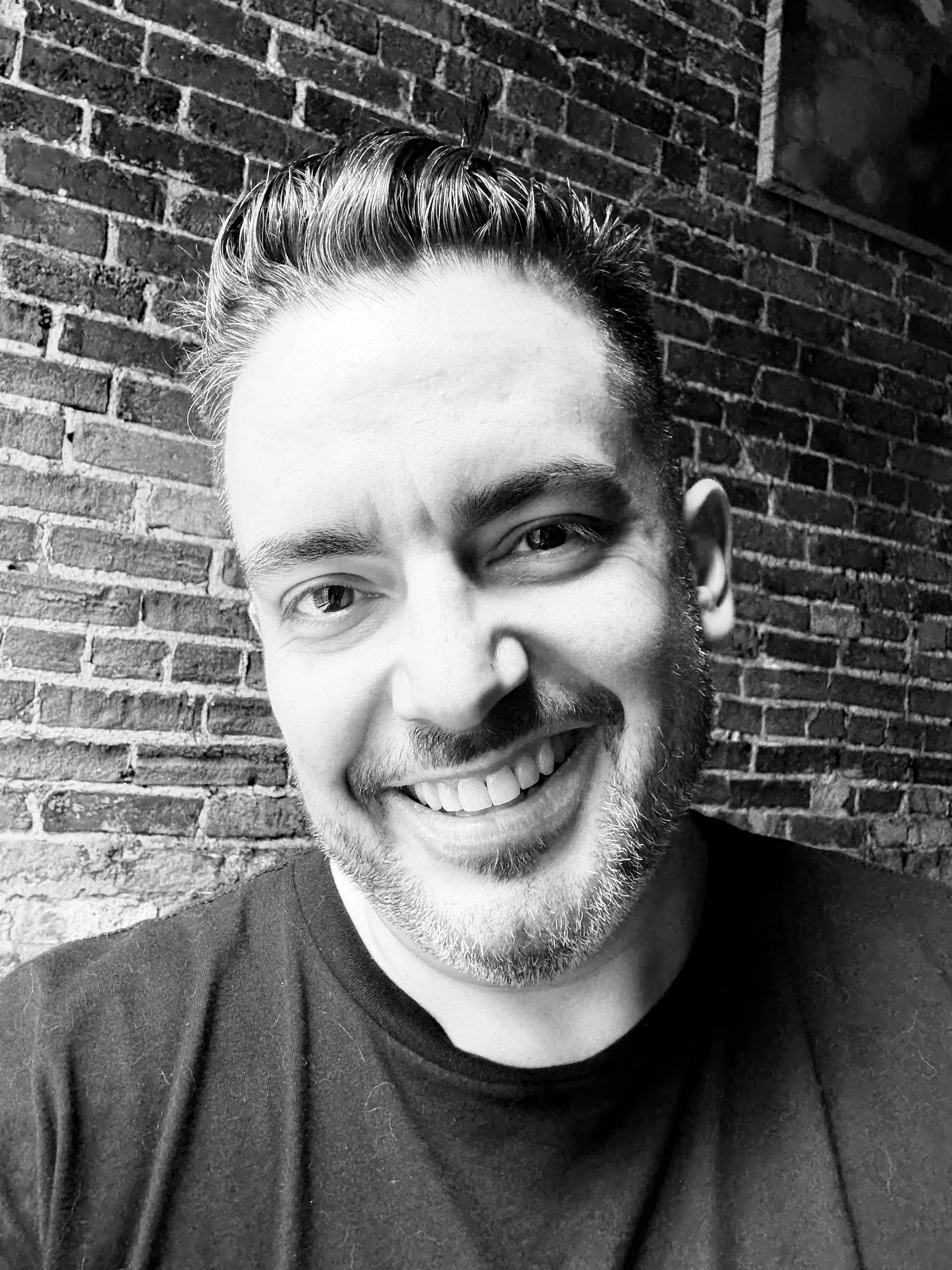 Black and white close-up portrait of a smiling man with short hair and a beard, standing in front of a brick wall.