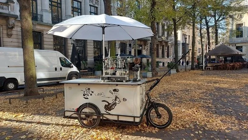 A mobile coffee or ice cream cart on a city street with fallen autumn leaves, has a large white umbrella and graffiti-style artwork on its side, parked near trees and buildings.
