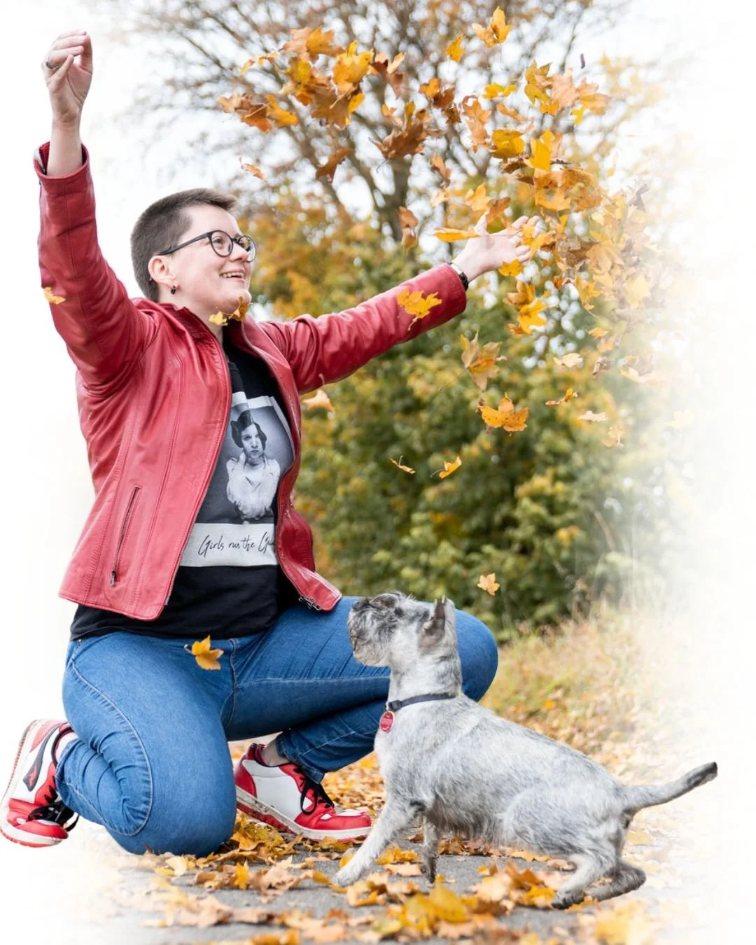 Eine Frau mit Gläsern in roter Lederjacke, die auf Knien im Herbstlaub steht und mit einem Lächeln Herbstblätter in die Luft wirft, kombiniert mit einem Dalmatiner-Hund, der auf einem Gehweg sitzt, umgeben von buntem Herbstlaub und Bäumen im Hintergrund.