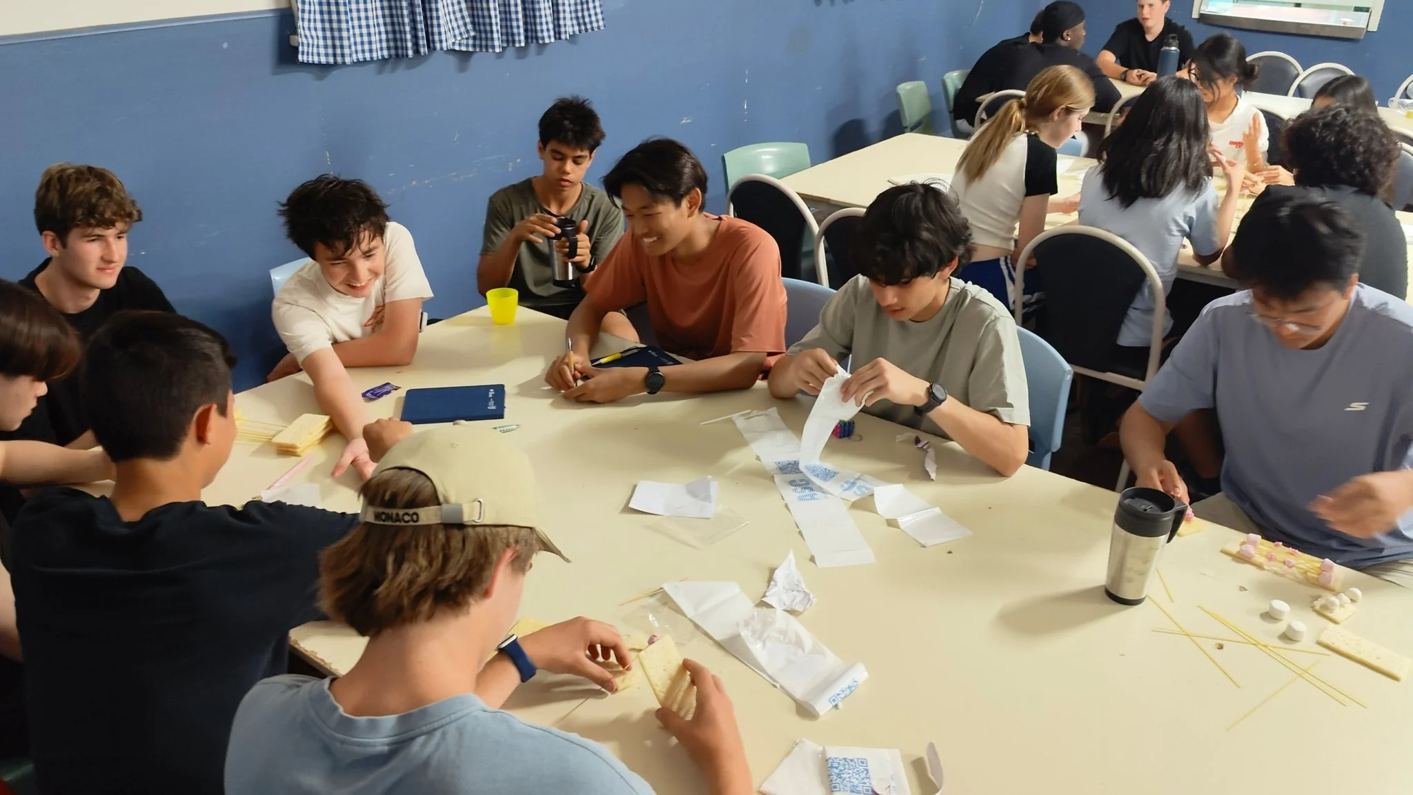 A group of young people gathered around tables working on a craft project with paper, pasta, and other materials in a classroom or community center setting.