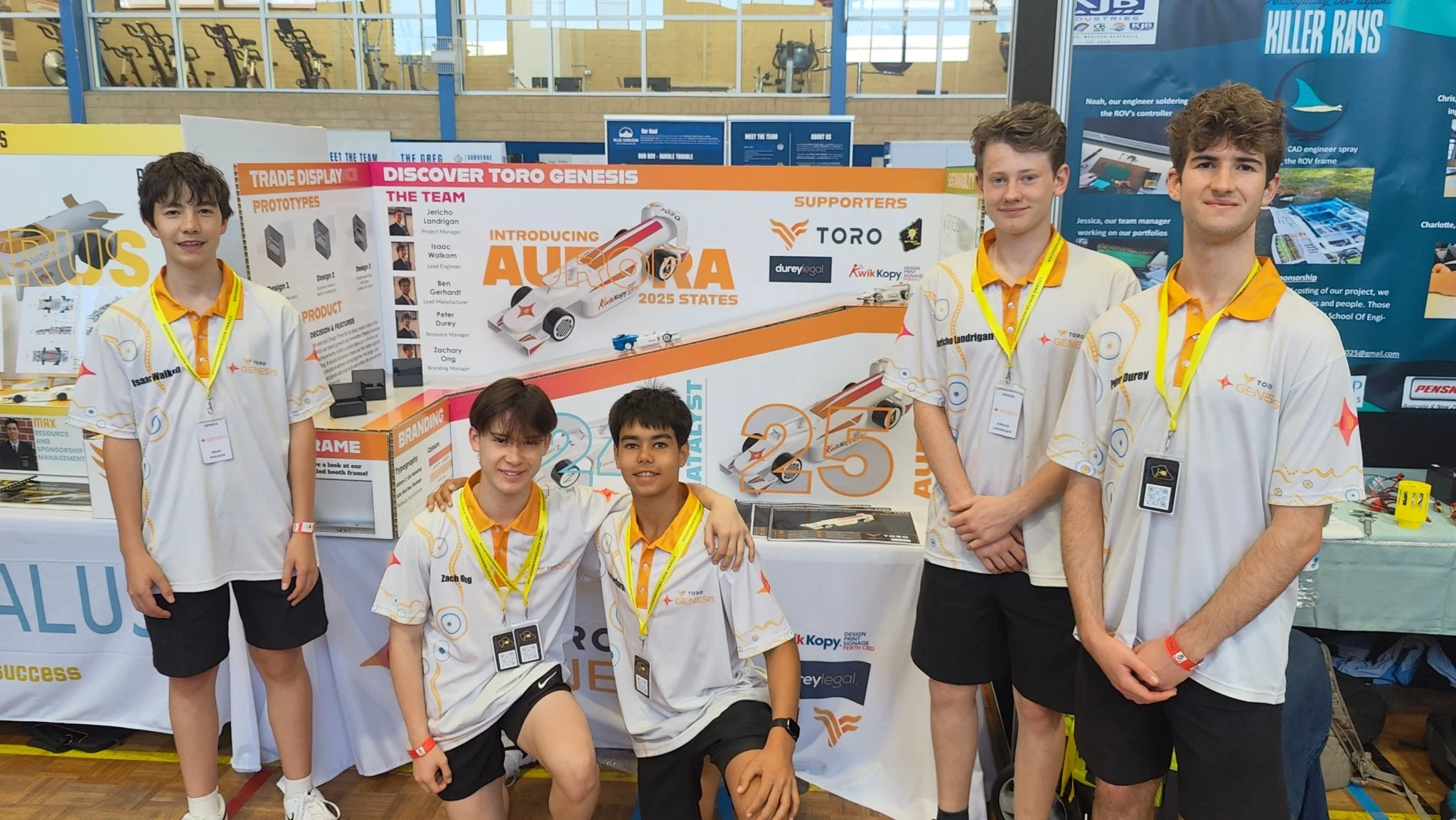 Five young boys wearing white team shirts with yellow accents standing in front of a science project display about autonomous vehicles at a science fair.