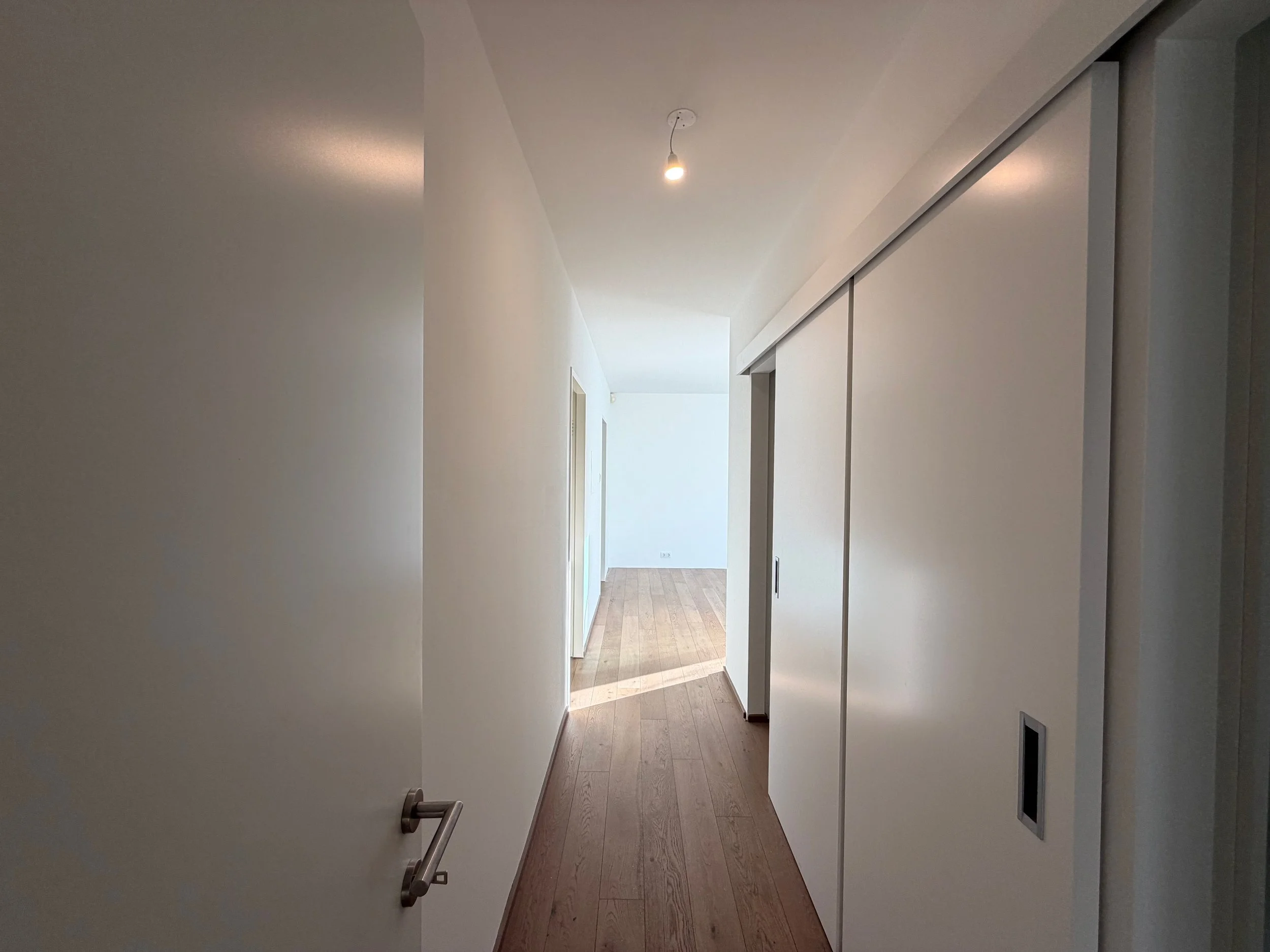 Weißer Flur mit Holzfußboden, geschlossene weiße Schiebetüren auf der rechten Seite, sichtbare Deckenleuchte, helles natürliches Licht im Hintergrund.