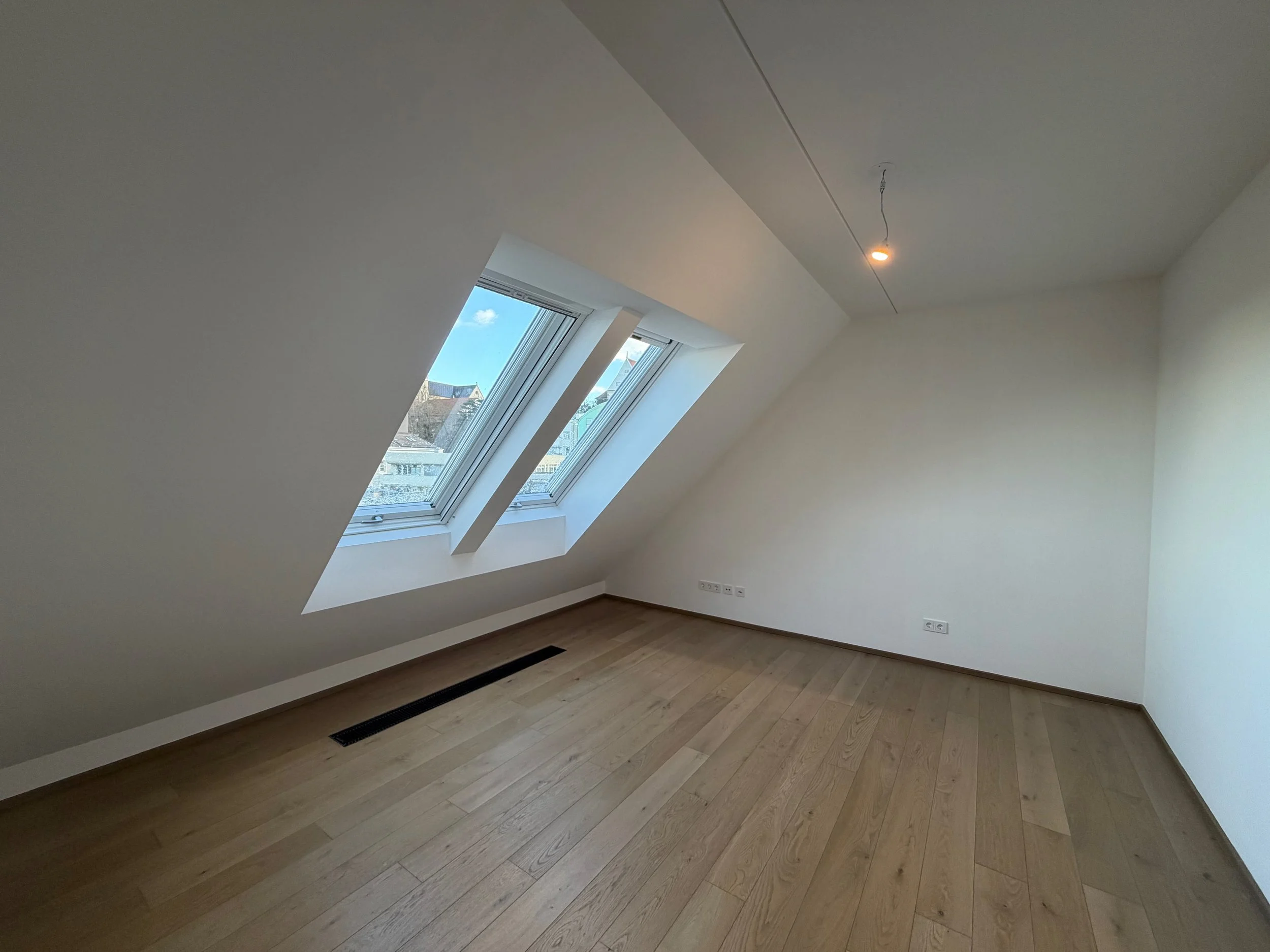Leeres Zimmer mit Dachfenstern, weißen Wänden, Holzboden und einer einzelnen Deckenleuchte, Blick nach draußen auf städtische Gebäude.