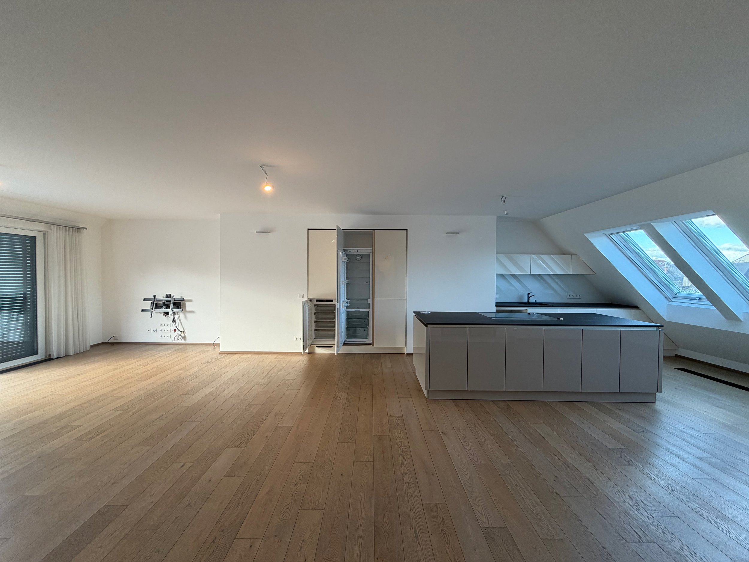 Leeres, modernes Wohnzimmer mit offener Küche, Holzfußboden, großen Fenstern, Balkon und weißen Wänden.