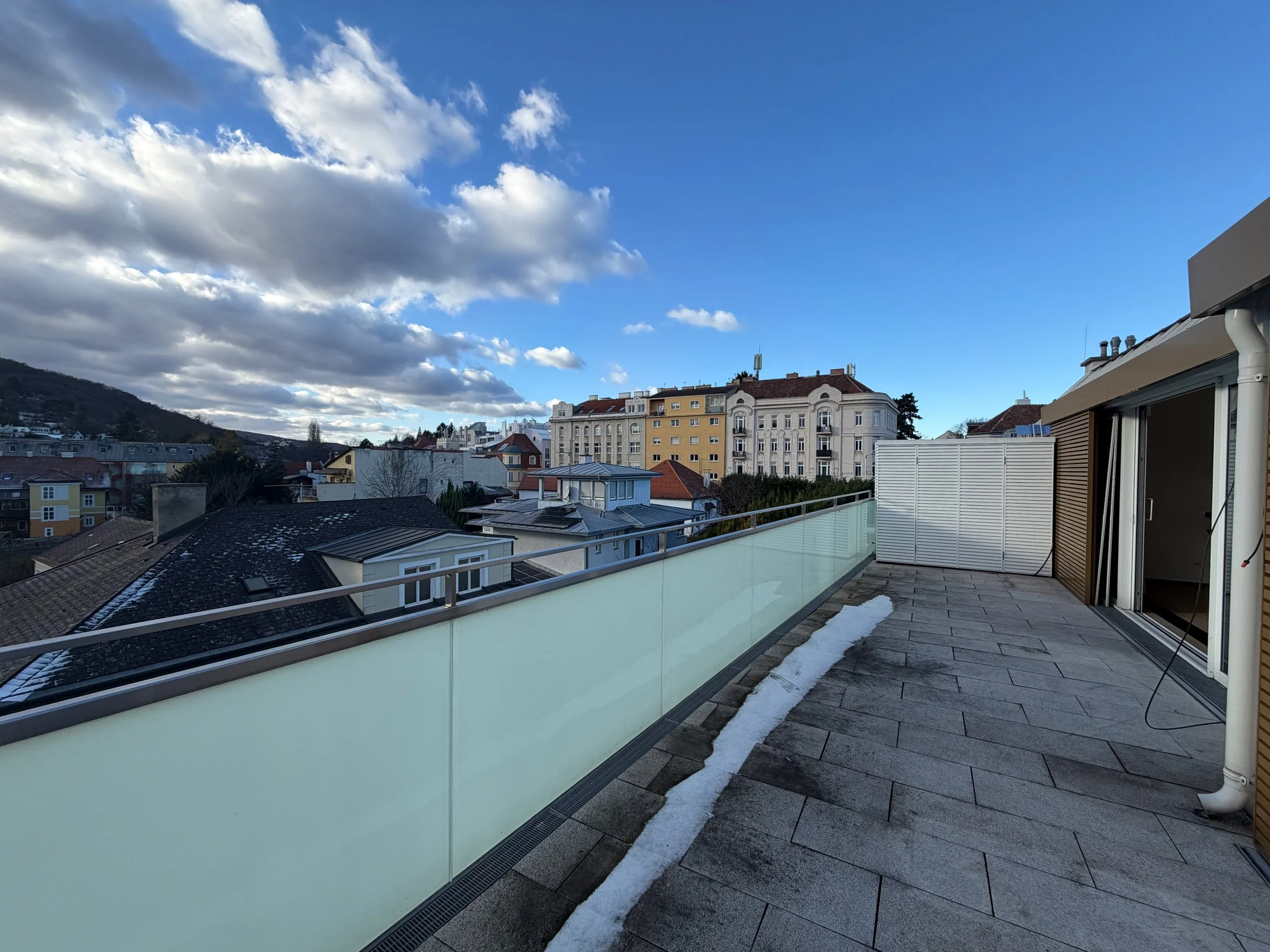 Balkon mit Glasgeländer, teilweise Schneereste auf dem Boden, umgeben von Stadtgebäuden und Bergen bei sonnigem Himmel mit Wolken.