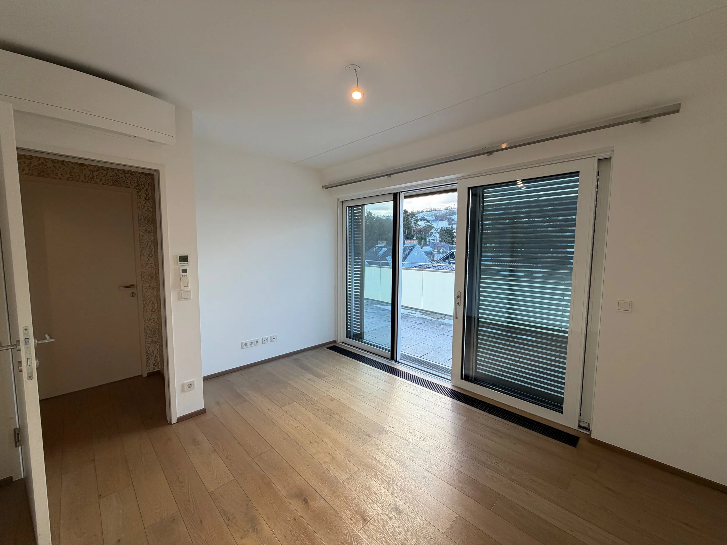 Leeres Wohnzimmer mit Holzboden, großen Glas-Schiebetüren zum Balkon, weißen Wänden und Deckenleuchte.