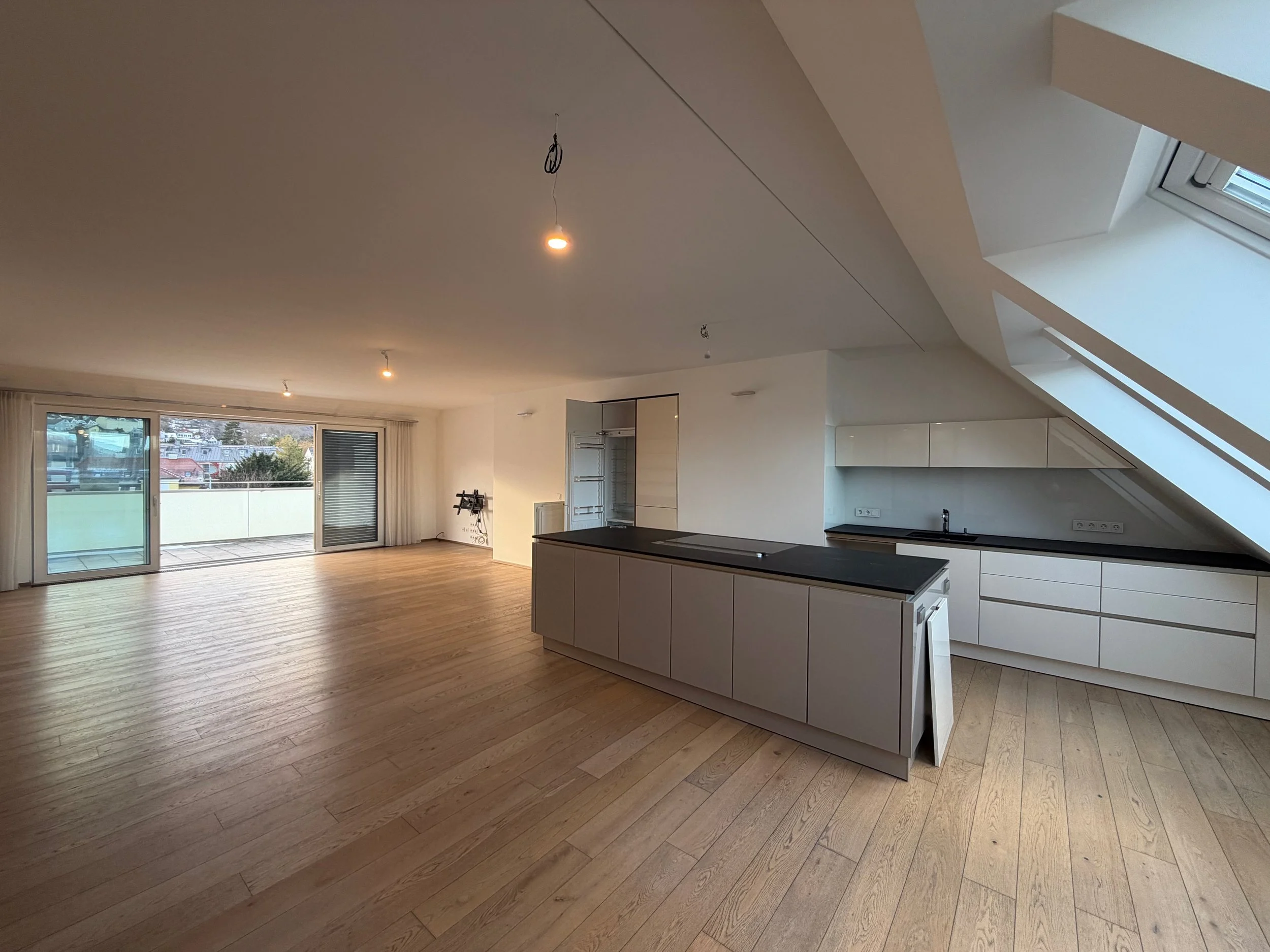 Leeres Wohnzimmer mit Küchenbereich, Holzboden, großen Fenstern, oder Balkon mit Stadtansicht, und Dachfensterelement, keine Möbel oder Dekorationen sichtbar.