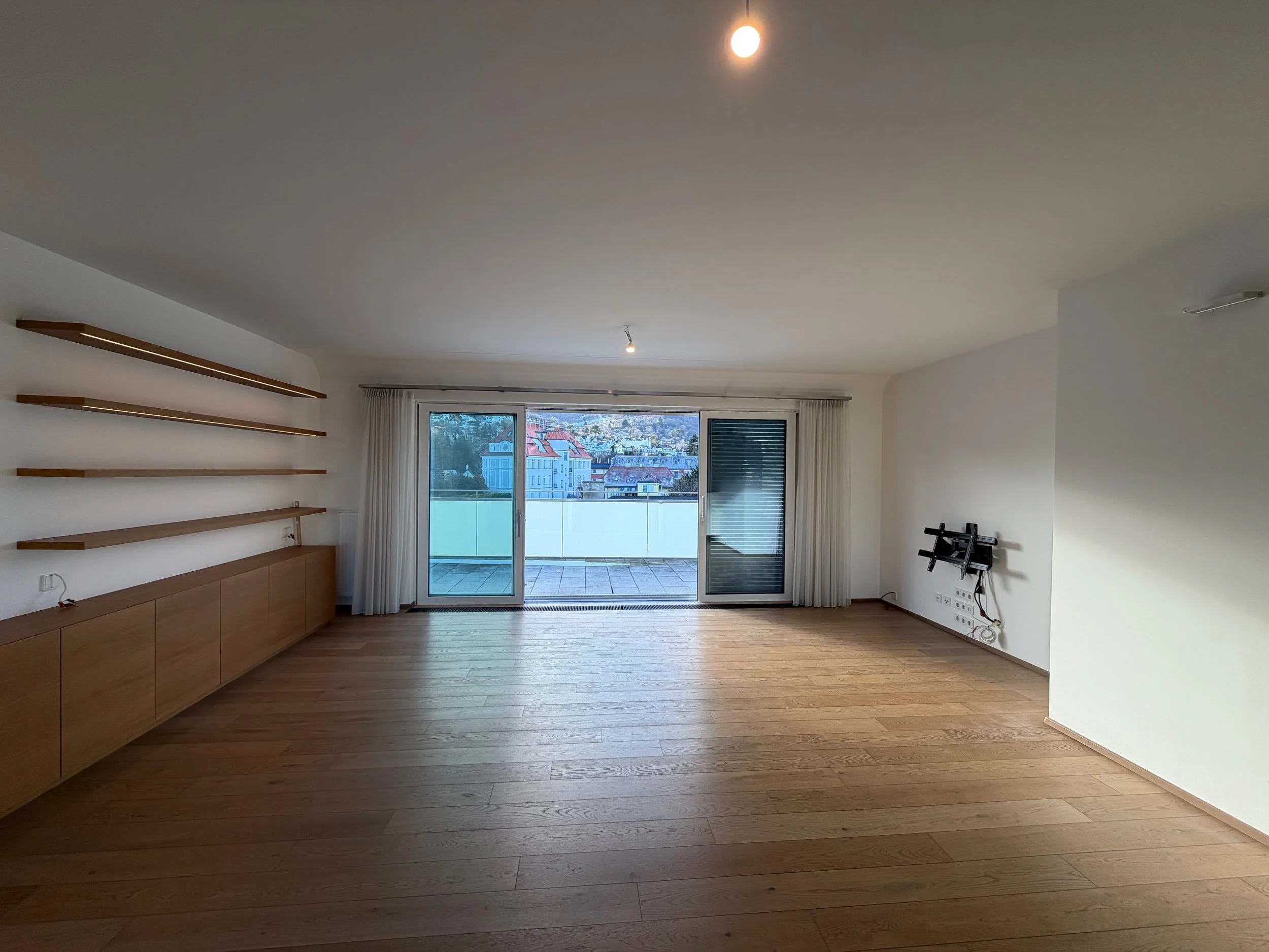 Leeres Wohnzimmer mit Holzboden, weißen Wänden, großen Fenstern mit Blick auf städtische Gebäude im Hintergrund, weiße Vorhänge, Holzregale an der linken Wand, und einer Wand mit einem nicht installierten Fernseher oder Bildschirmanschlüssen.