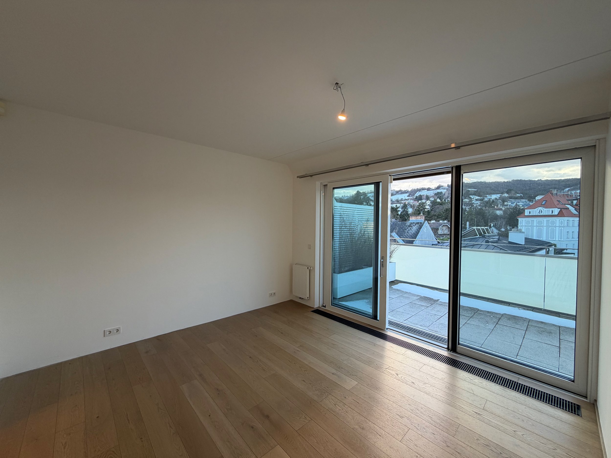 Leeres Zimmer mit großen Glas-Türen, die zu einem Balkon führen, und Blick auf eine Stadt mit Hügeln im Hintergrund.