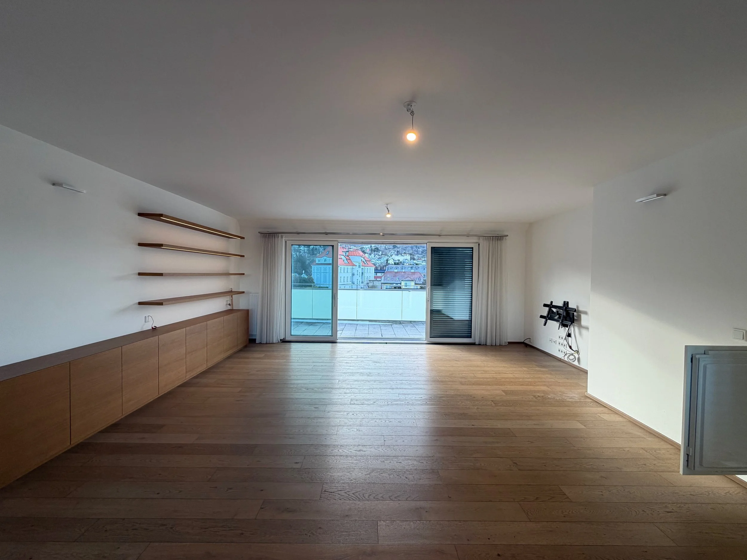 Leeres Wohnzimmer mit Holzboden, weißen Wänden, an der Wand sind leere Regale und ein langes Holzregal. Es gibt große Fenster und eine Balkontür, die nach draußen führen. Die Vorhänge sind offen, und es sind zwei unbeleuchtete Lampen an der Decke.