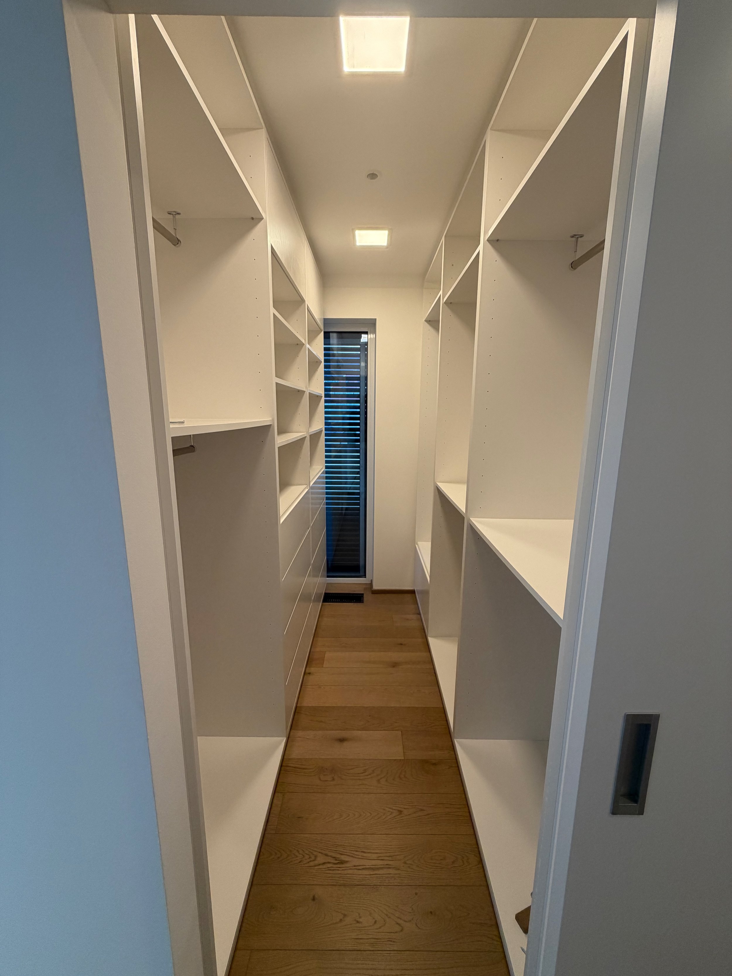 Ein begehbarer Kleiderschrank mit weißen Einbauschränken auf beiden Seiten, Holzboden und einem Fenster an der Rückwand.