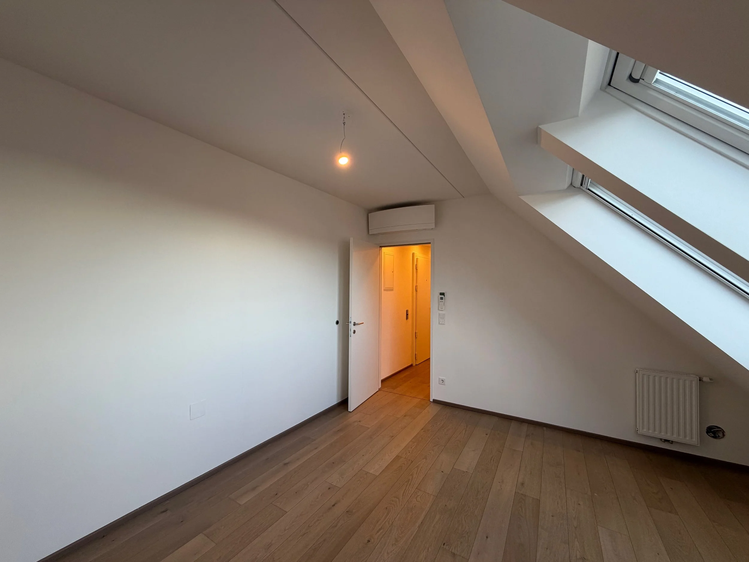 Leeres Zimmer mit Holzboden, weißen Wänden, einer Dachschräge mit großen Dachfenstern, einem offenen Tür, einer Deckenleuchte und einer Heizkörper unter den Fenstern.